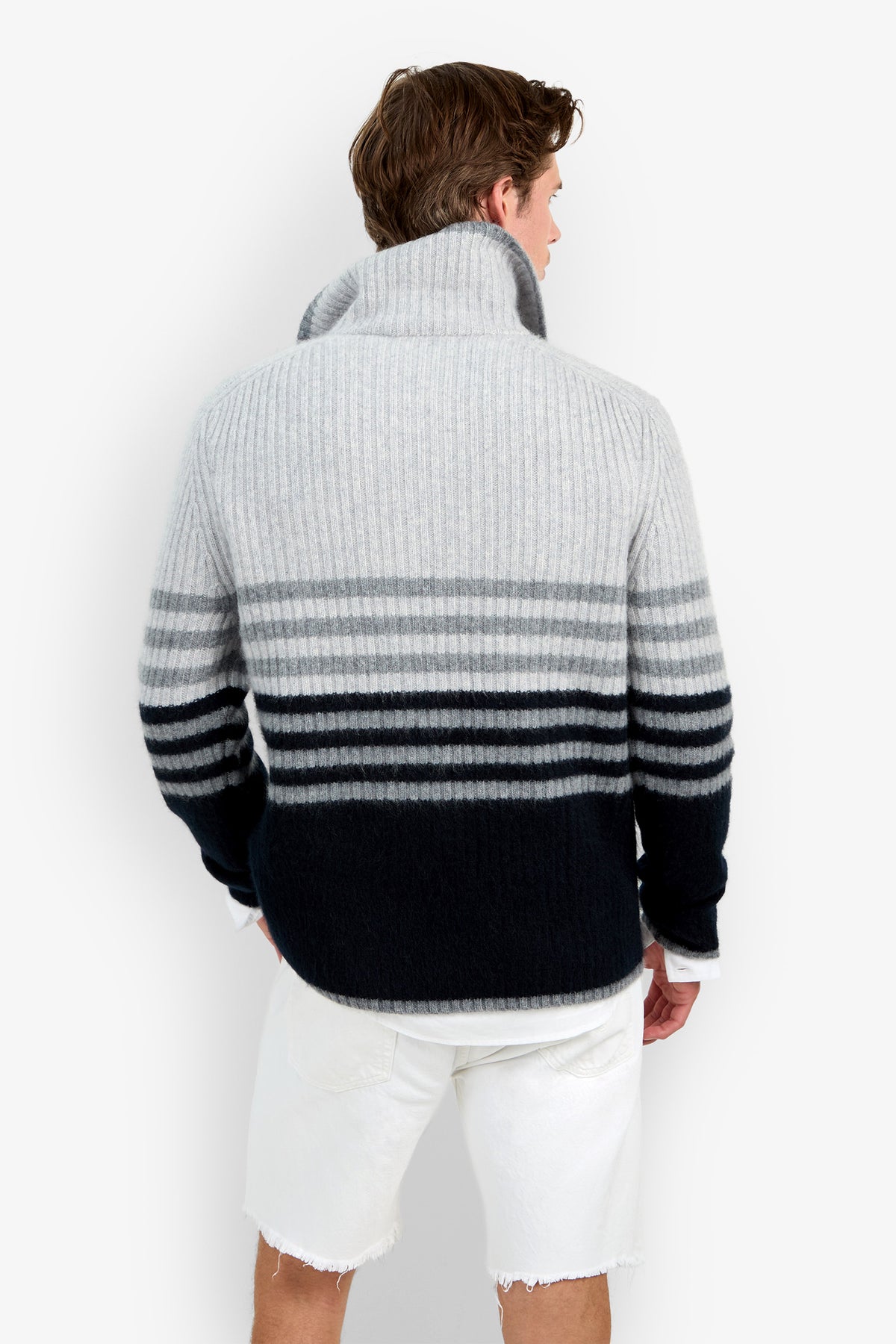 A man facing away wears the HEMMINGWAY QUARTER ZIP PULLOVER—a thick, ribbed, striped sweater in shades of gray and black—paired with frayed white shorts.