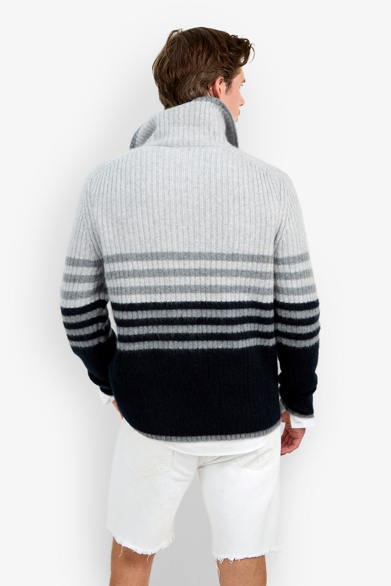 A man facing away wears the HEMMINGWAY QUARTER ZIP PULLOVER—a thick, ribbed, striped sweater in shades of gray and black—paired with frayed white shorts.
