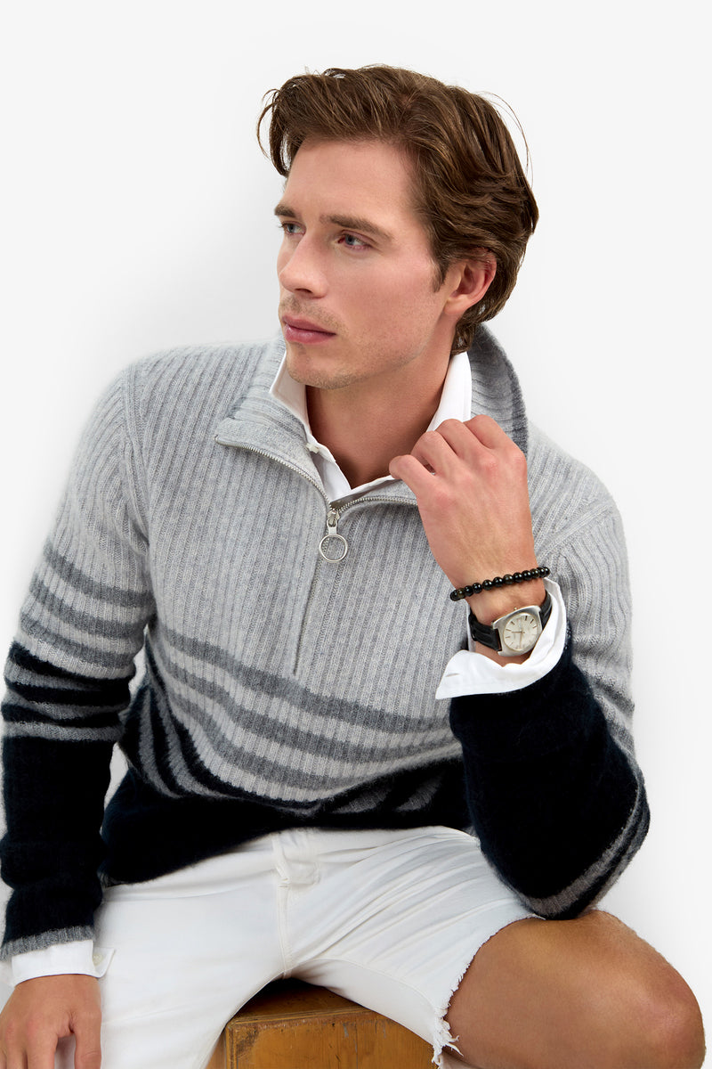 Wearing the HEMMINGWAY QUARTER ZIP PULLOVER, a man with light brown hair sits on a wooden stool in white shorts and a white collared shirt, looking to the side with his hand near his collar.