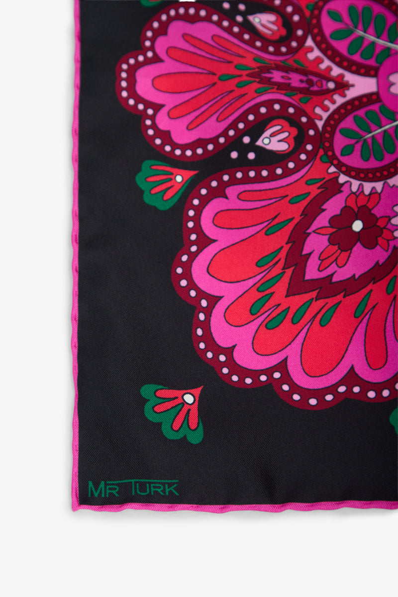 A close-up of the Sautoir Silk Pocket Square showcases black fabric adorned with vibrant pink, red, and green floral patterns. The corner features a label with “MR TURK” in green text.