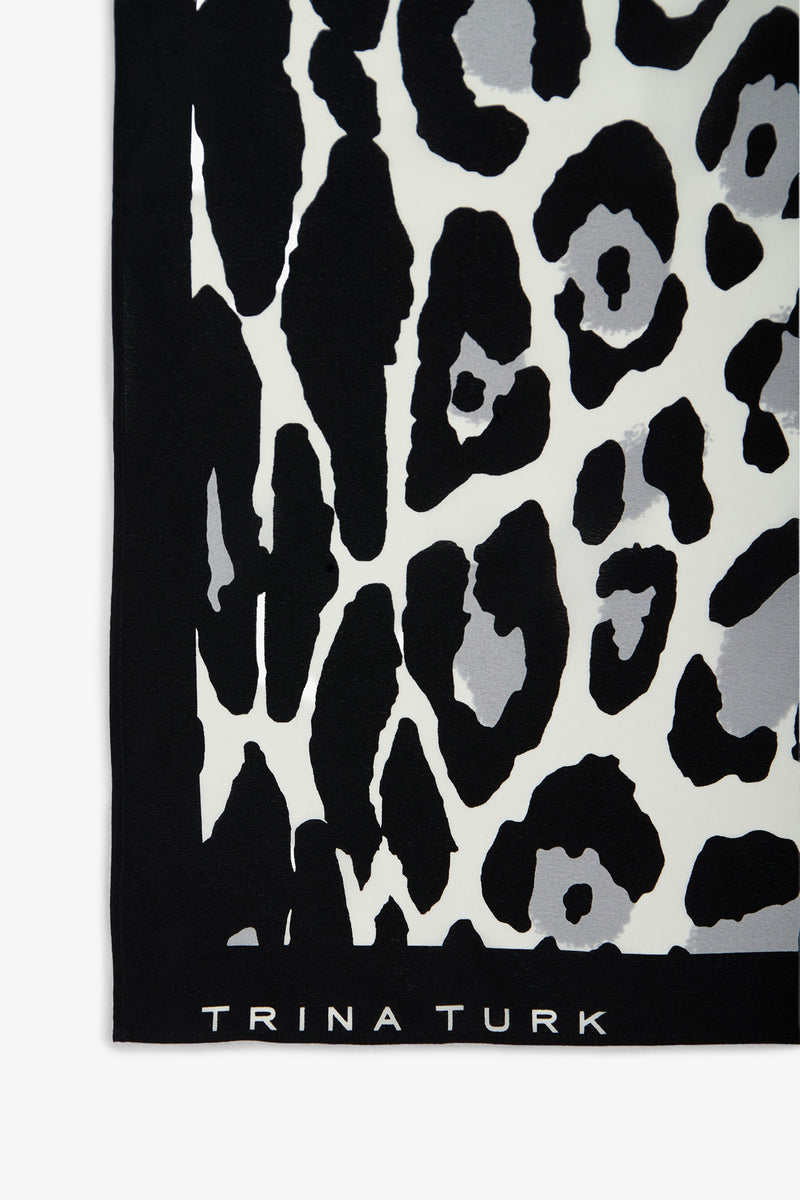 A section of the Snow Leopard Silk Scarf featuring a black, white, and gray leopard print pattern, with “TRINA TURK” displayed in white along the bottom edge.