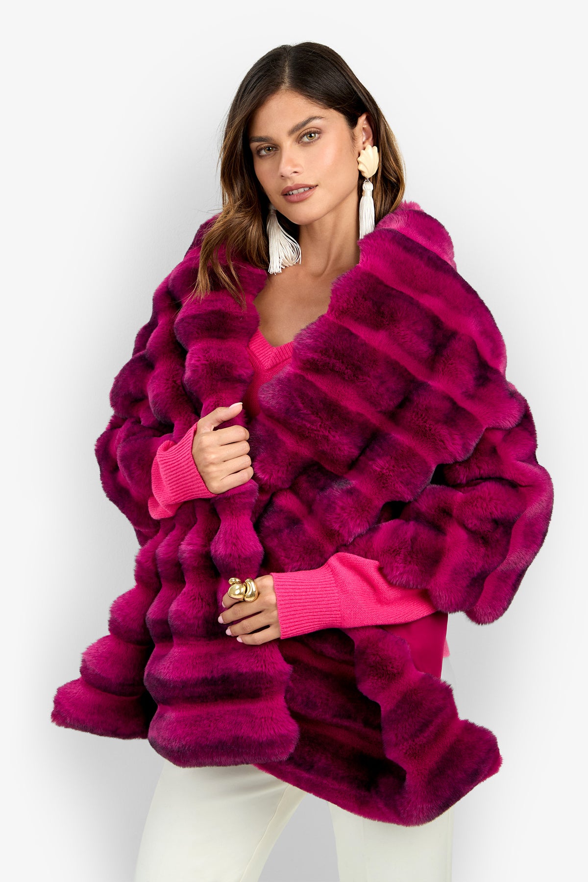 A woman wearing the Mikel Fur Stole in TORMALINA, styled with large white tassel earrings and white pants, poses against a plain light background.