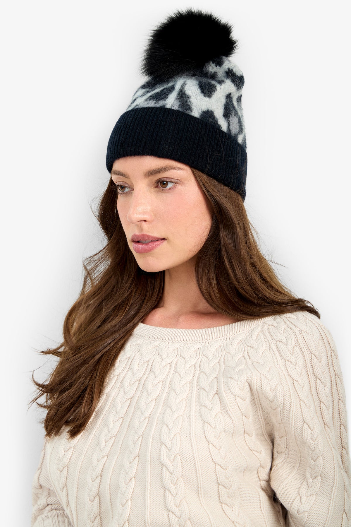 A person wears a CARDUCCI LEOPARD HAT, a cashmere beanie featuring a black and gray leopard pattern, a solid black ribbed cuff, and a black pom-pom.
