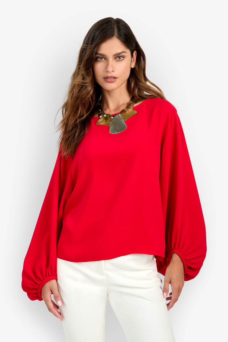 A woman wearing the URSA 2 TOP in bright red polyester with gathered long sleeves and a large gold-toned statement necklace.