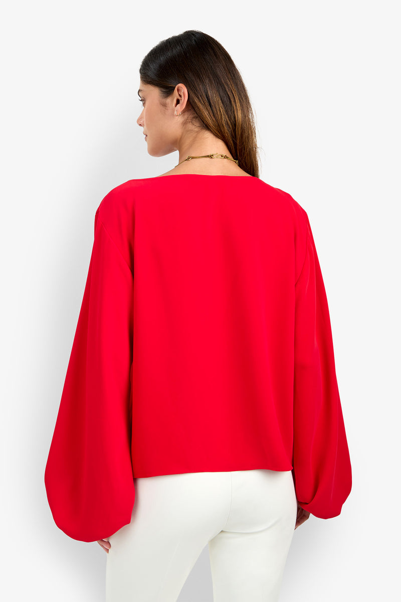 A woman seen from behind wearing the bright red, long-sleeved URSA 2 TOP made of polyester with a wide neckline and loose, billowy sleeves, paired with white trousers.