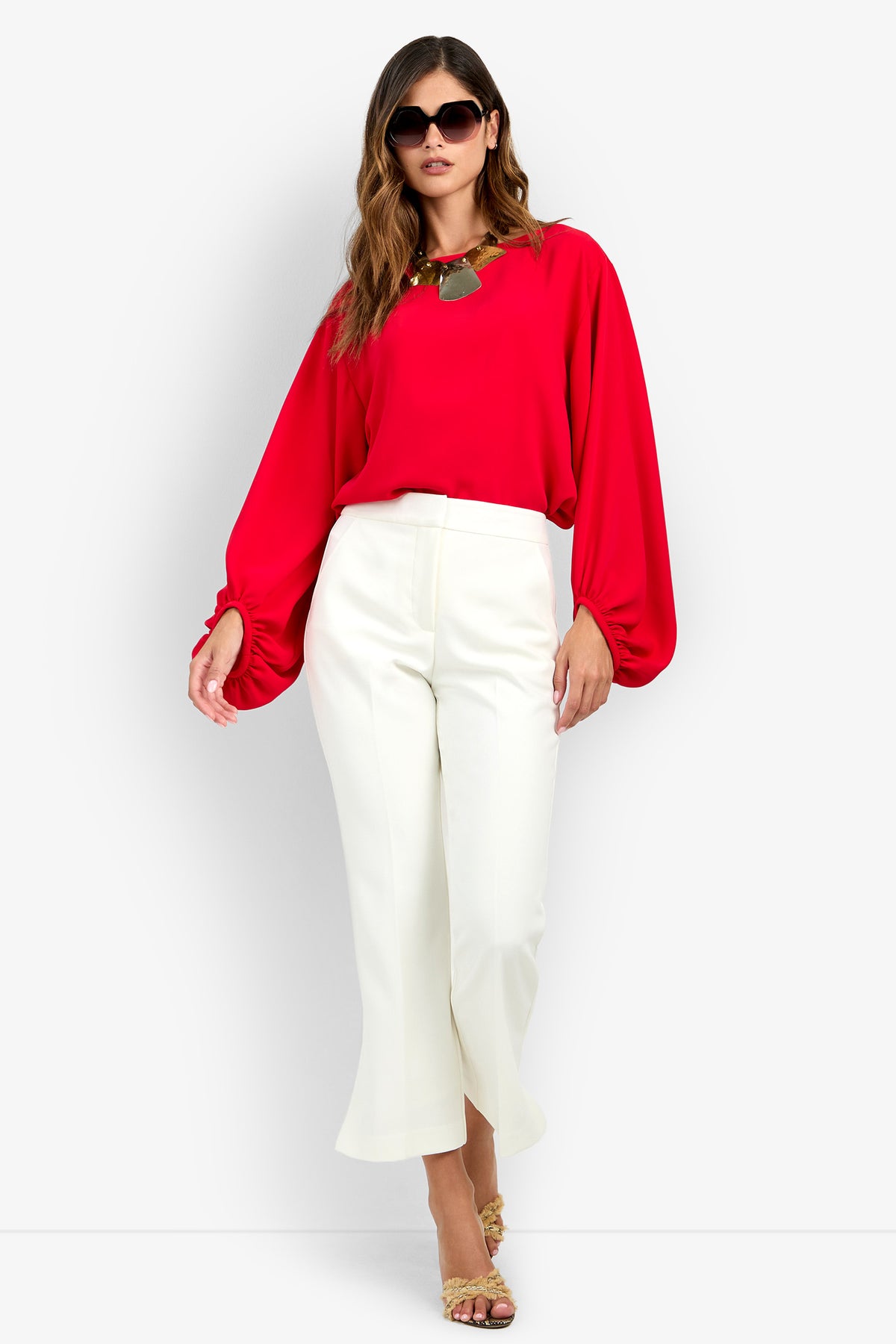 A woman wearing the red URSA 2 TOP with long sleeves and cinched cuffs, paired with white trousers and oversized sunglasses.