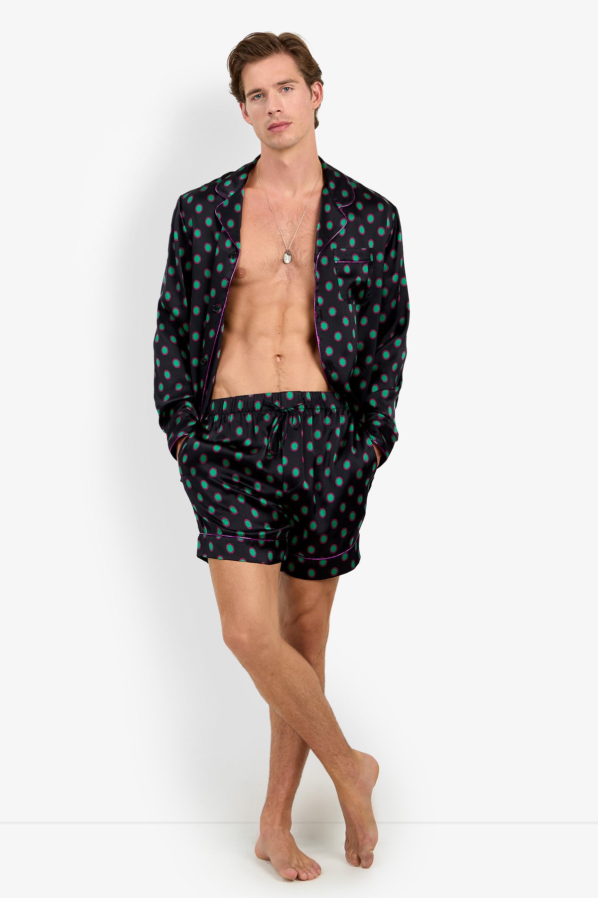 A man wearing a PJ TOP made of 100% silk with matching shorts, featuring a black pattern with green polka dots and purple piping.