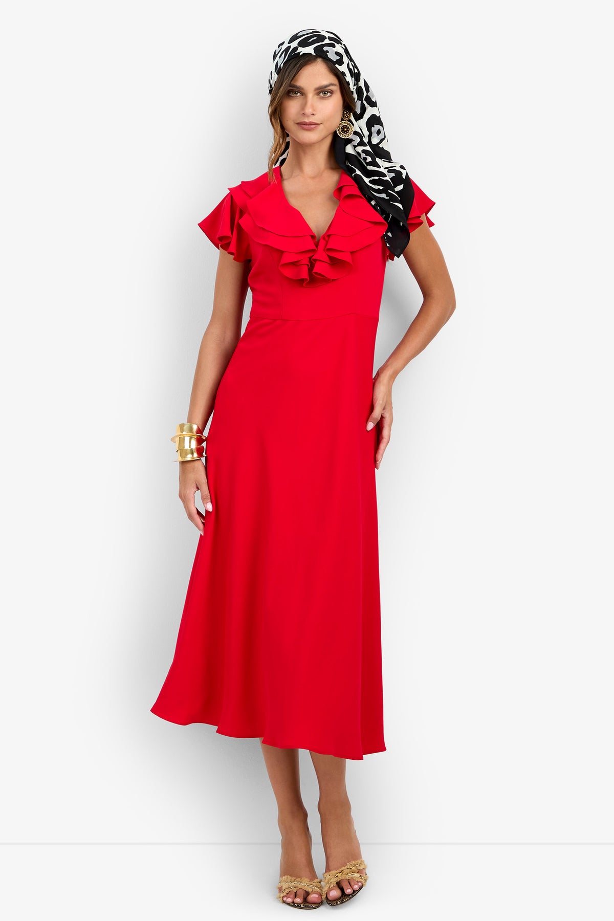 A woman wearing the red polyester PATRONS DRESS with ruffled cap sleeves and a V-neckline, styled with a black and white patterned headscarf and gold cuff bracelet.