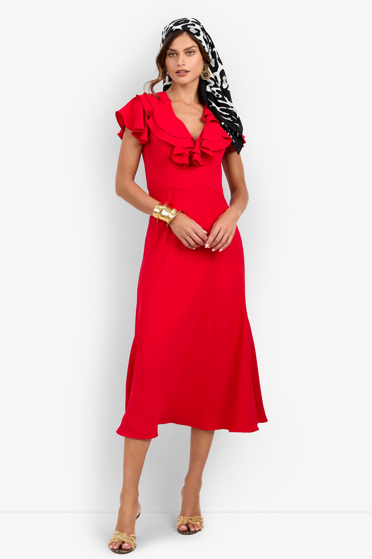 A woman wearing a red PATRONS DRESS with ruffled sleeves and a V-neckline, paired with a black and white patterned headscarf, gold bracelets, and sandals.
