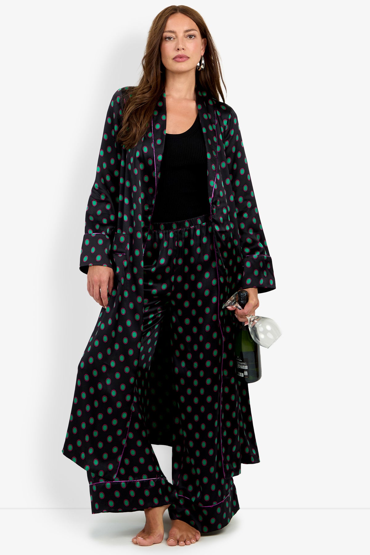 A woman wears an ADALYNN ROBE in 100% silk with matching wide-leg trousers, both featuring a black pattern with green and purple polka dots, while holding a bottle of sparkling wine and a glass.