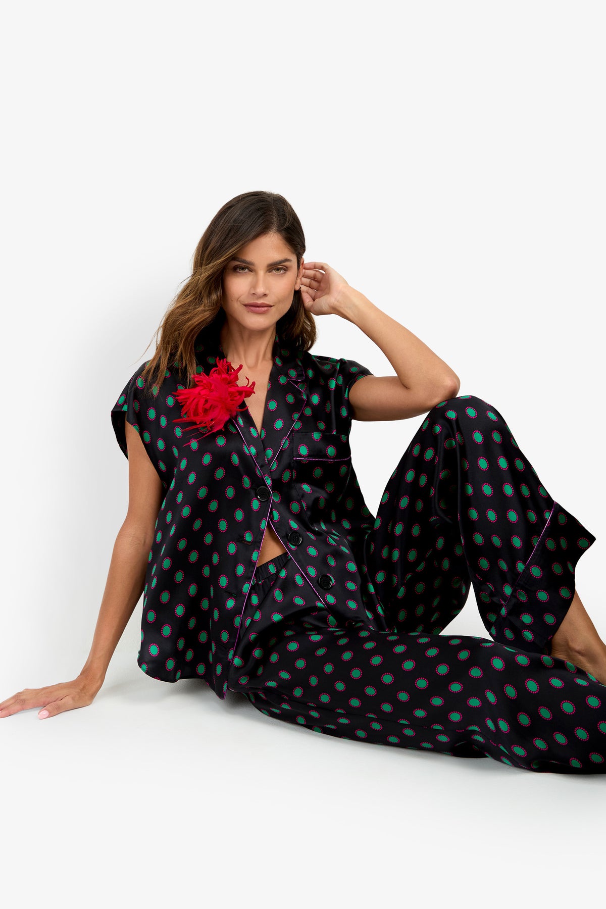 A woman sits on the floor wearing a pure silk matching black button-down shirt and LIARA PANT set with a green and pink circular pattern, featuring a red feather embellishment on the shirt’s lapel.