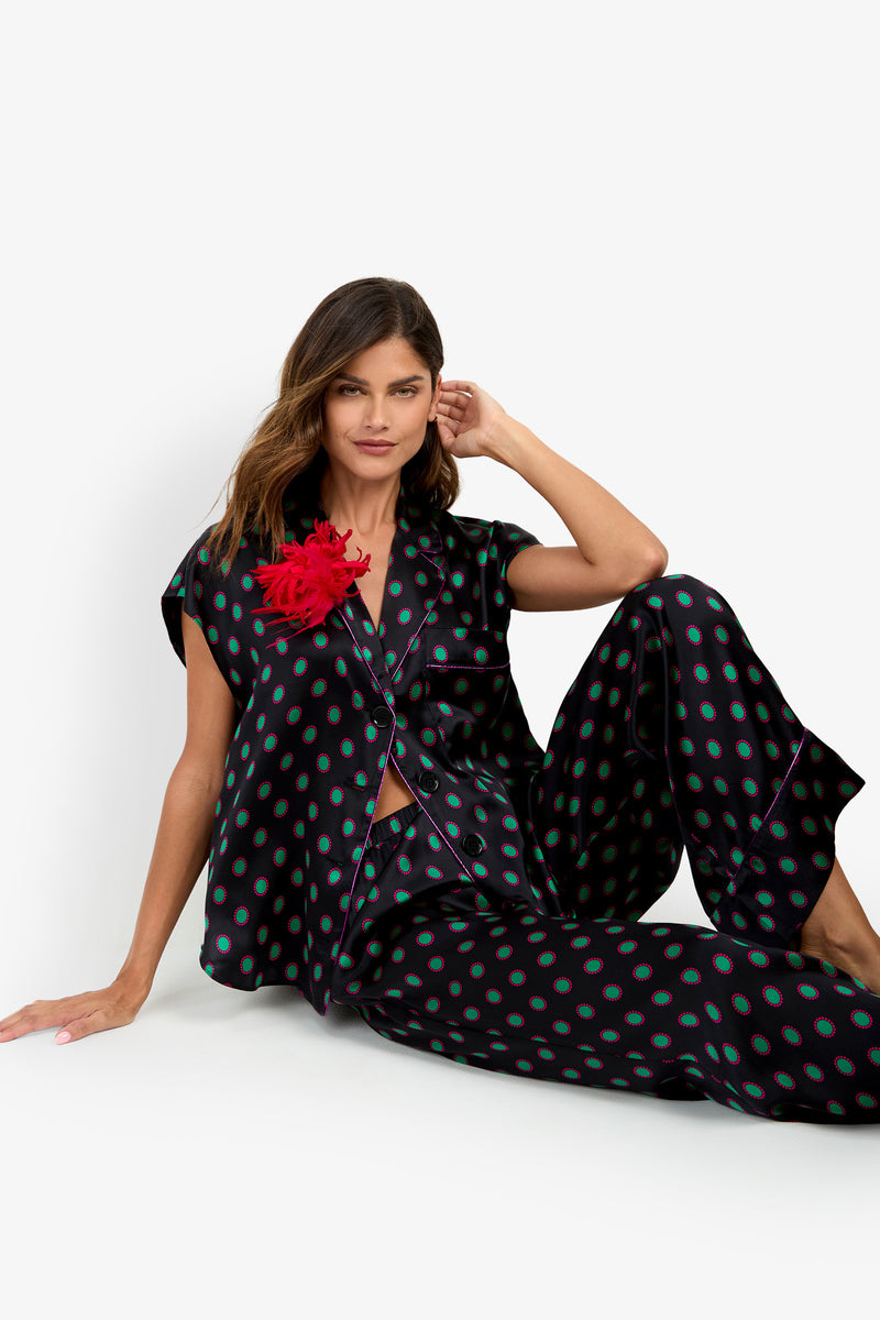 A woman sits on the floor wearing a pure silk matching black button-down shirt and LIARA PANT set with a green and pink circular pattern, featuring a red feather embellishment on the shirt’s lapel.