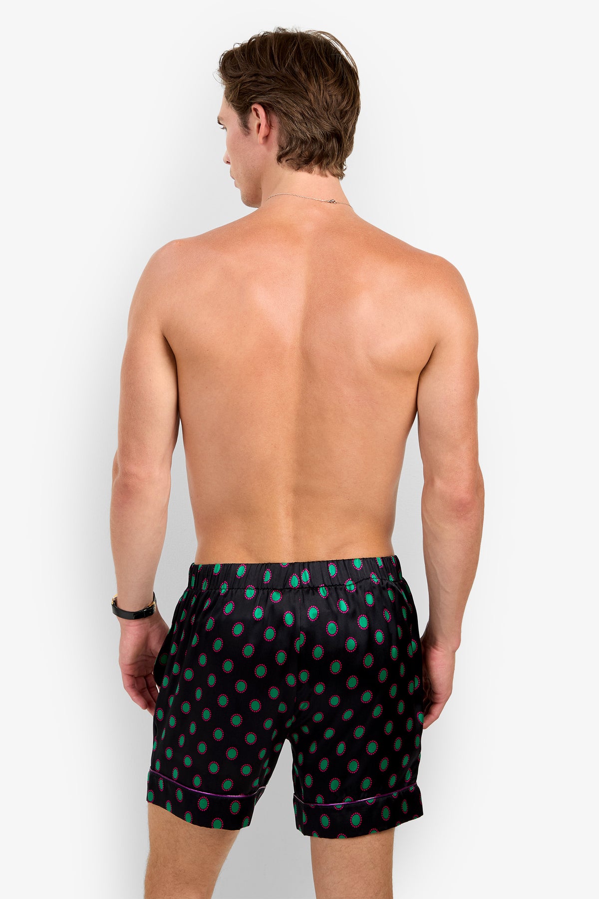 A shirtless man from behind wearing PJ SHORT black pajama shorts made of pure silk, featuring a repeating teal and red circular pattern and purple piping.
