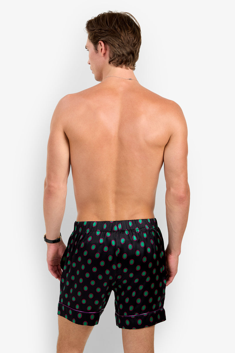 A shirtless man from behind wearing PJ SHORT black pajama shorts made of pure silk, featuring a repeating teal and red circular pattern and purple piping.