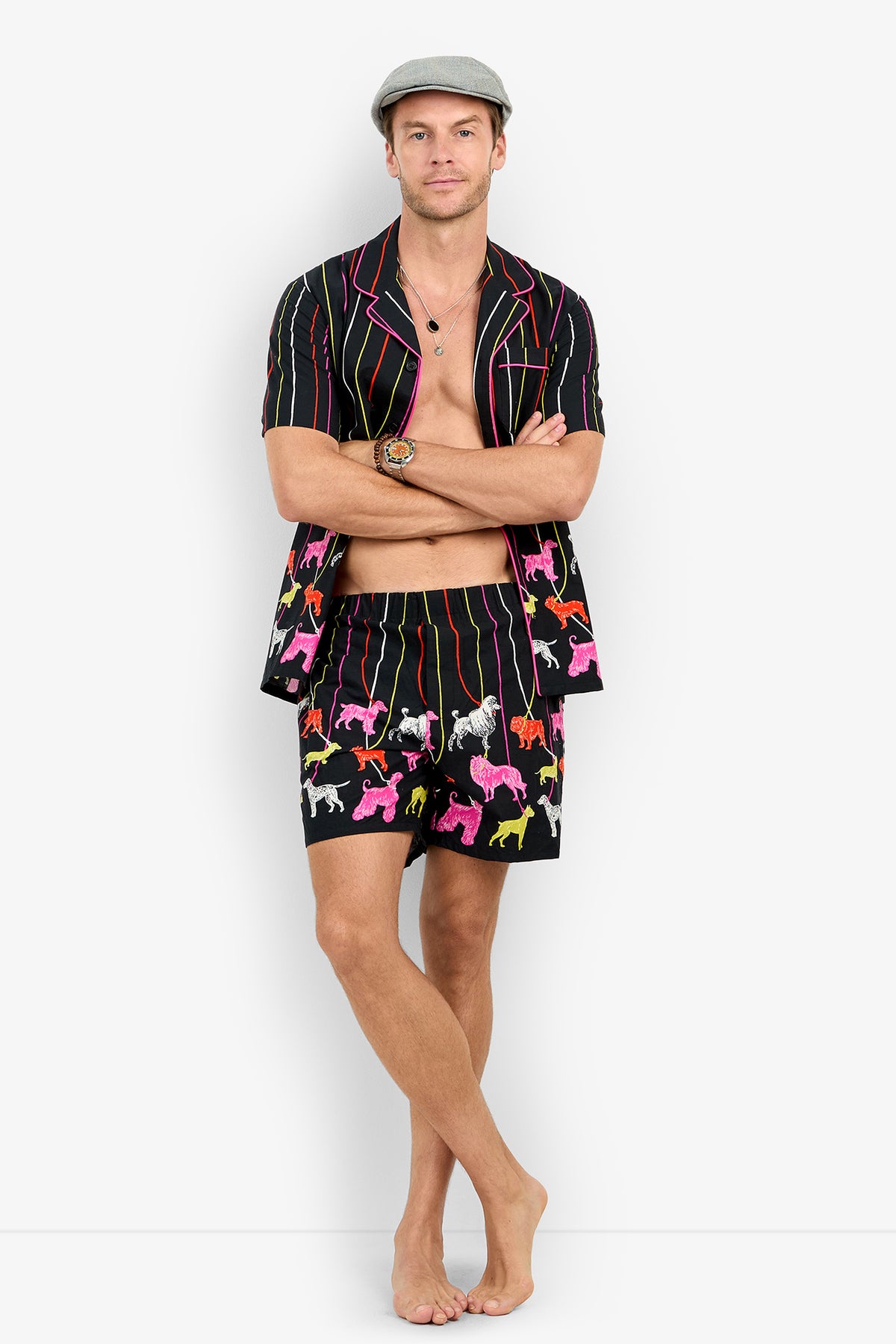 A man wearing a PJ SHORT set featuring a short-sleeved shirt and matching shorts with a pattern of colorful dogs and vertical stripes.