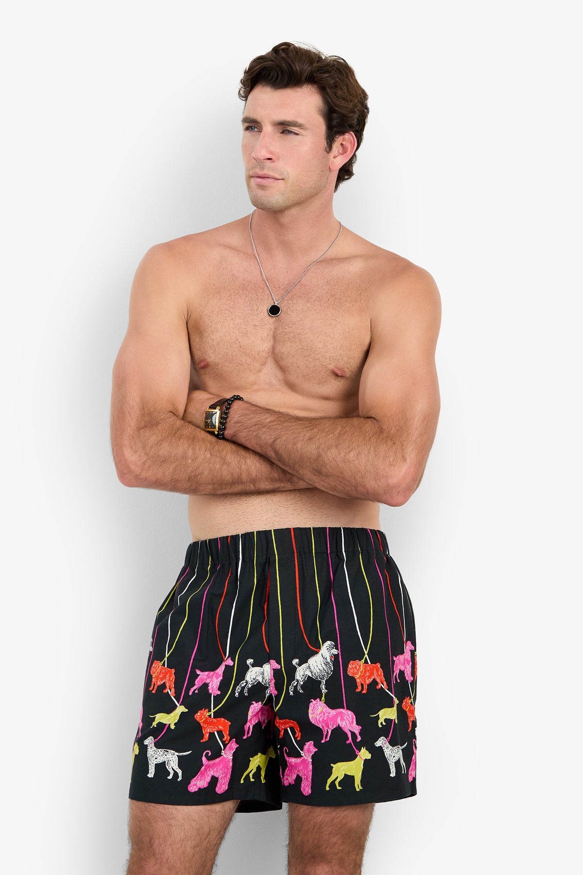 A shirtless man with his arms crossed wearing PJ SHORT black boxer shorts decorated with a colorful pattern of dogs on leashes.