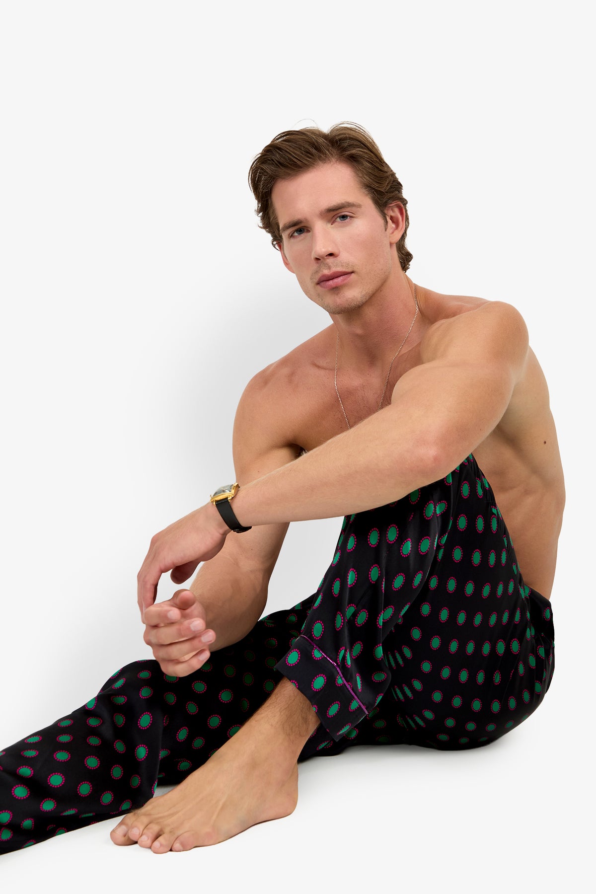 A shirtless man sits wearing PJ PANT silk pajama pants featuring a repeating pattern of green and pink circles.