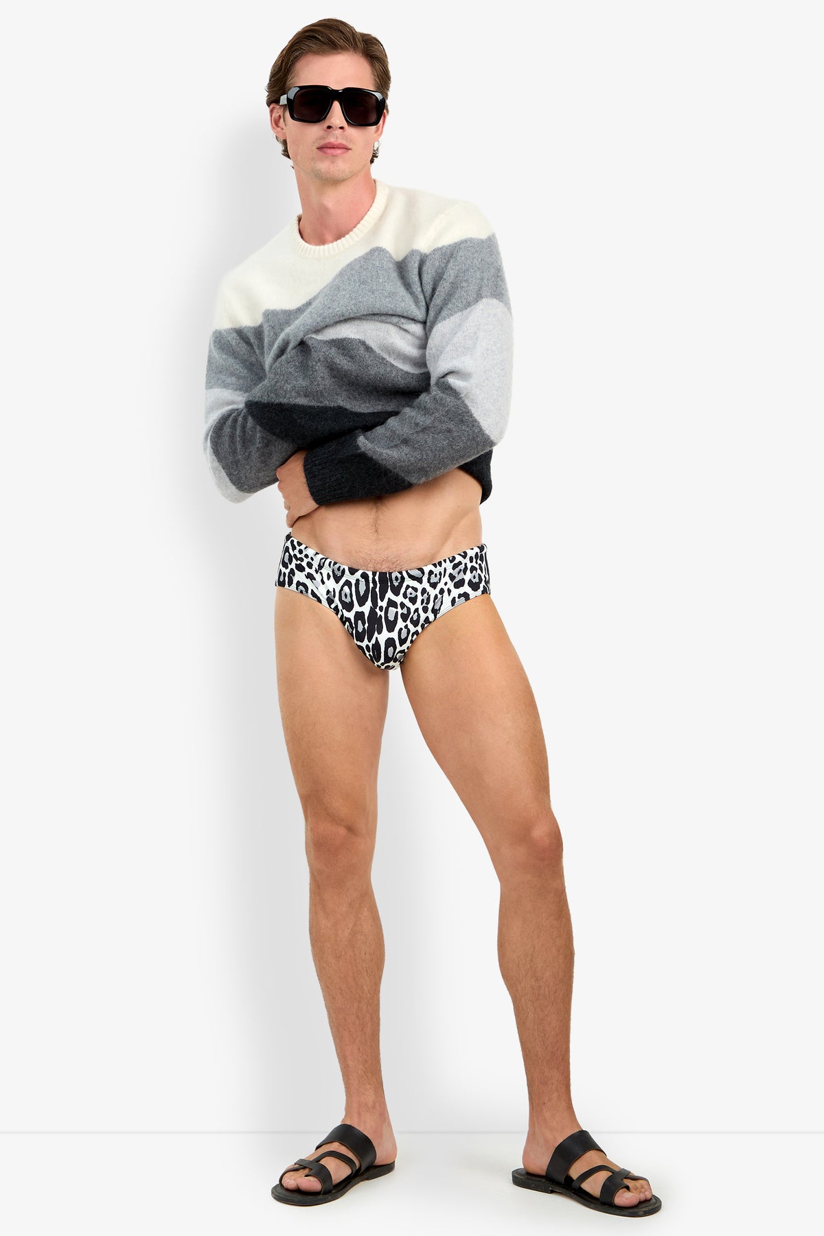 A man wearing black sunglasses, a grey and white cashmere color-block sweater, and a BANNING leopard-print silk swimsuit, standing in black sandals against a white background.