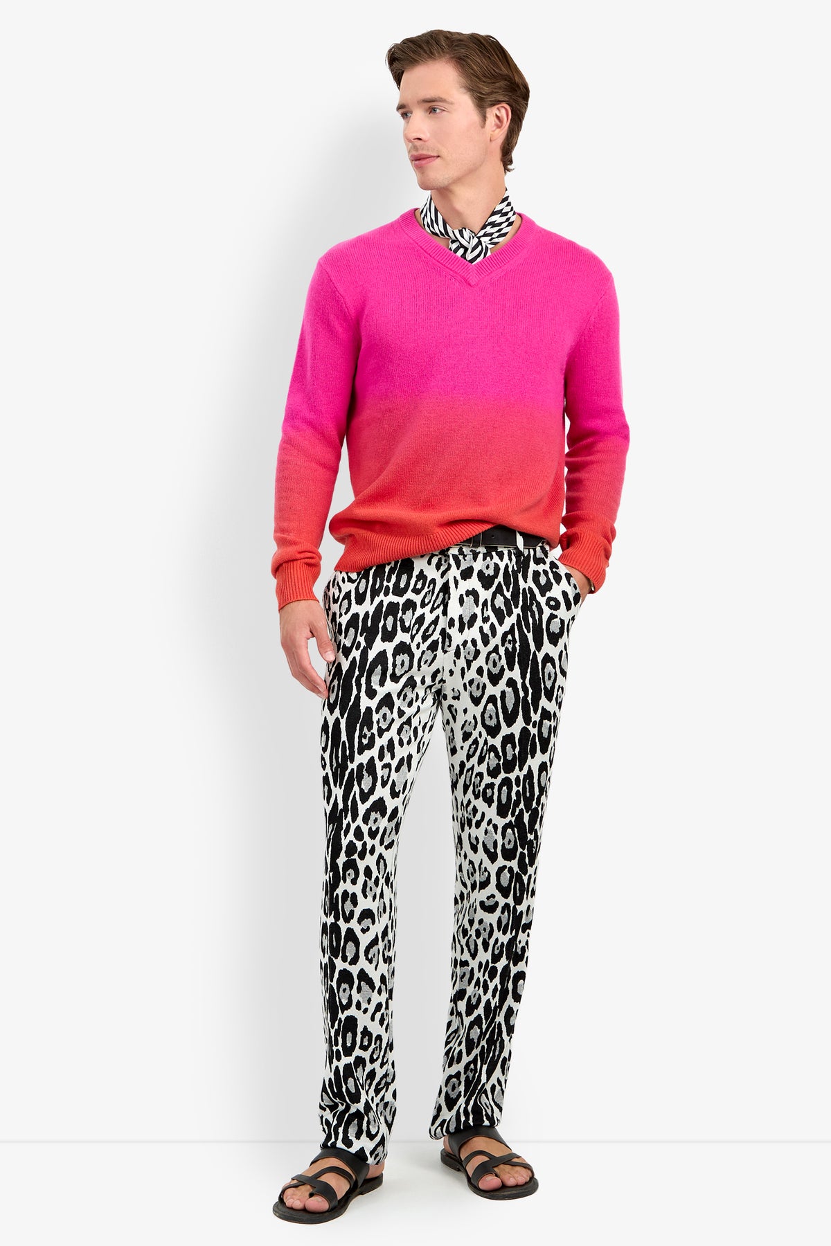 A man wearing a TIMOTHY OMBRE pink-to-red cashmere v-neck sweater, black and white leopard print pants, a patterned neck scarf, and black sandals.