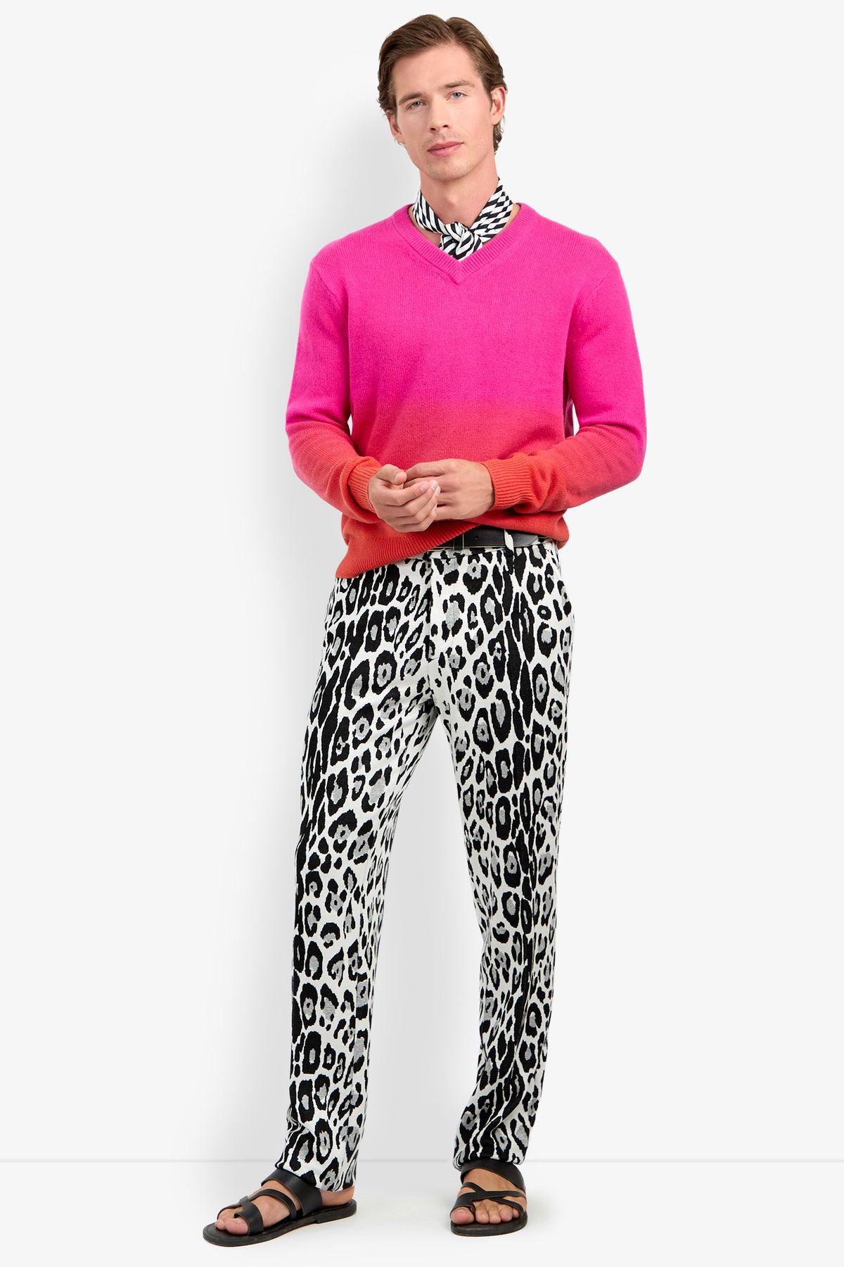 A man wearing a TIMOTHY OMBRE pink and orange v-neck cashmere sweater, a black and white patterned neck scarf, and leopard print trousers with black sandals.