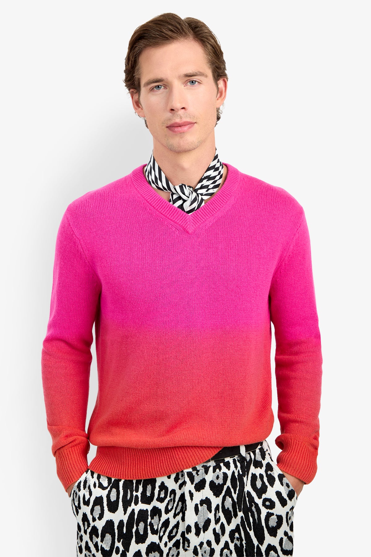 A man wears the TIMOTHY OMBRE pink-to-orange cashmere V-neck sweater, a black and white patterned neck scarf, and black and white leopard print trousers.
