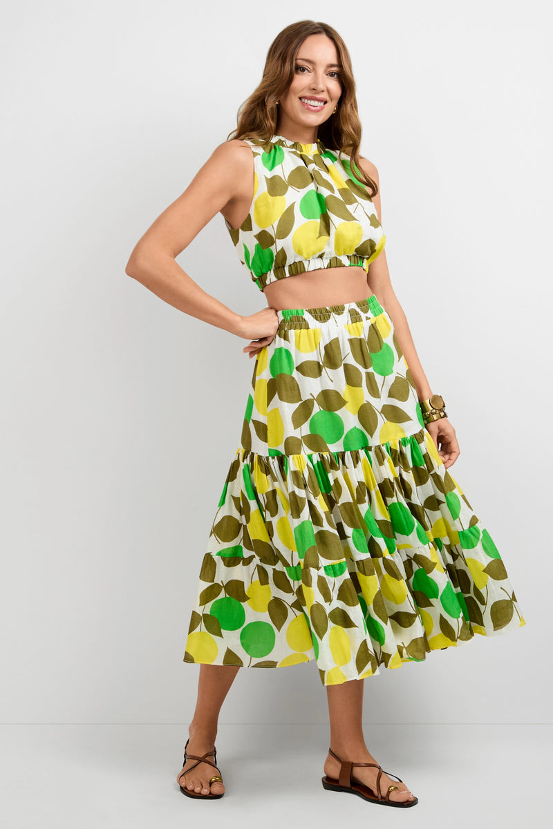 A woman smiles while wearing the Kalila Crop Top—a sleeveless, collared crop top featuring a green, yellow, and olive leaf print—paired with a matching tiered skirt against a plain white background. She accessorizes with sandals and a bracelet.