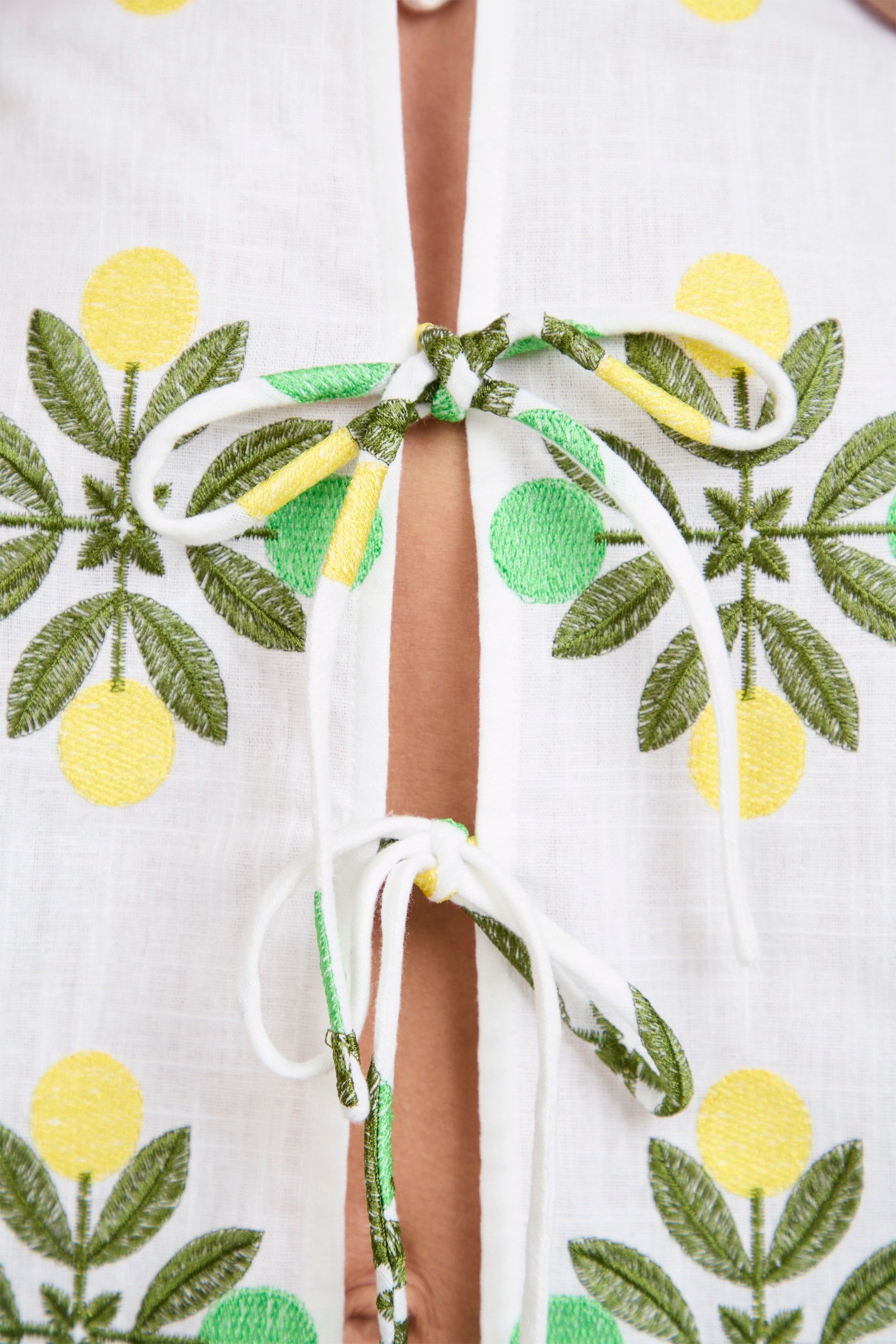 Close-up of the Salina Tie-Front Top, a white garment with green and yellow embroidered lemons and leaves, featuring two white fabric ties fastened in bows at the front that partially reveal skin underneath.