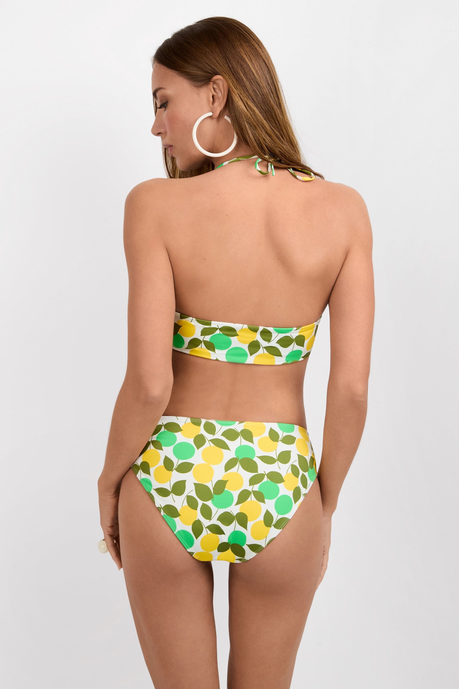 Woman with long brown hair stands with her back to the camera, wearing the Kammie Swim Top - Limone, a retro-patterned yellow, green, and white bandeau bikini top paired with matching high-waisted bottoms and large hoop earrings.