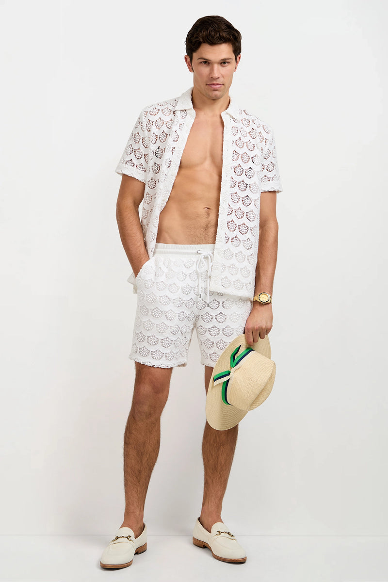A man stands against a white background wearing the Branson Crochet Lace Shirt, white drawstring shorts, white loafers, a gold watch, and holding a straw hat with a green stripe.