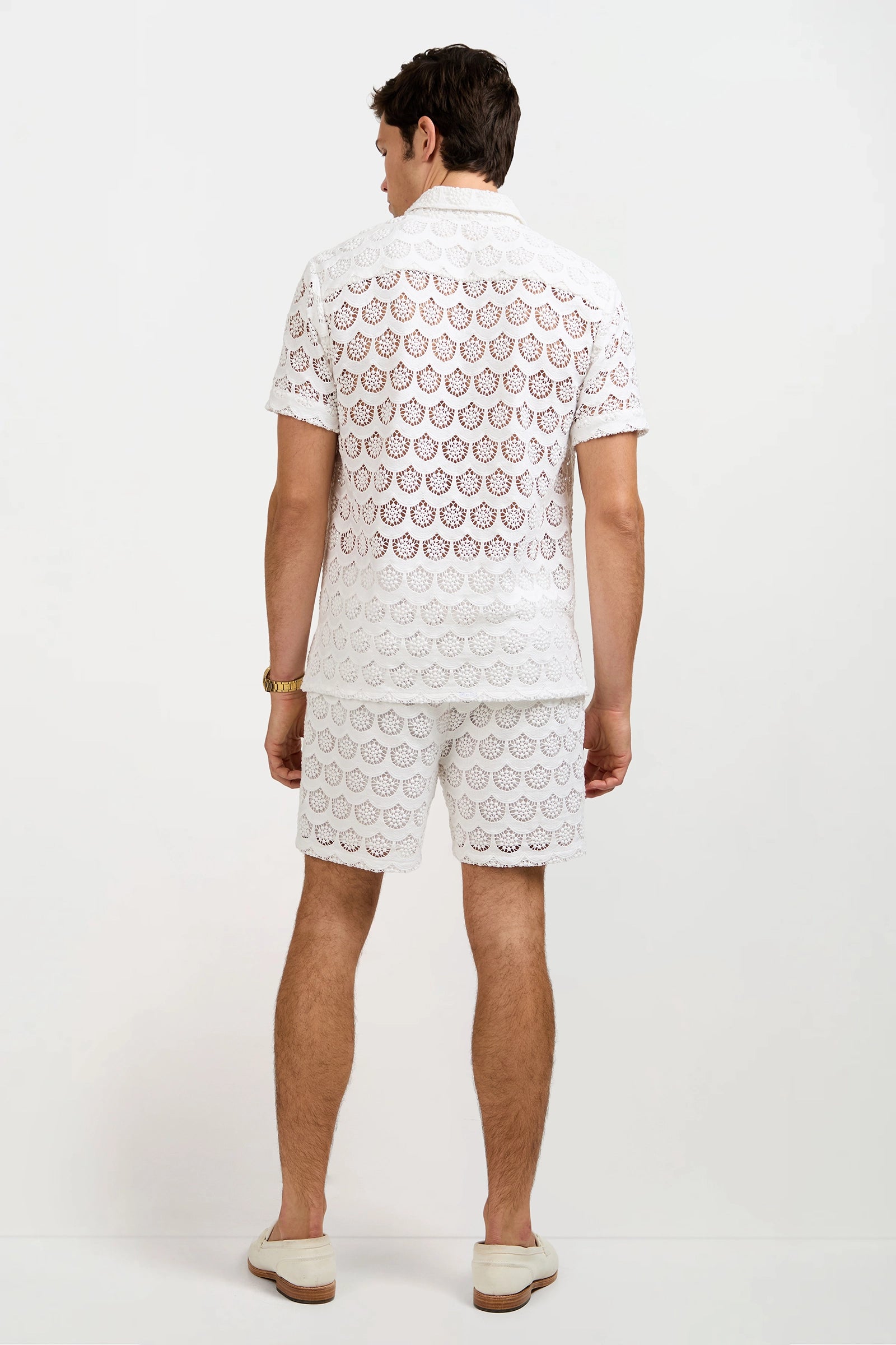 A man with light skin and short dark hair faces away, wearing the Shoreline Crochet Lace Short as part of a matching white lace shirt and shorts set with circular patterns, paired with white slip-on shoes. The background is plain white.
