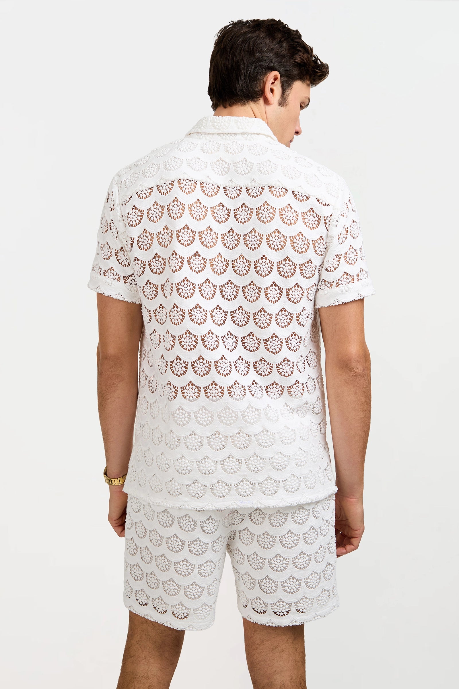 A man stands with his back to the camera, wearing the Branson Crochet Lace Shirt and matching shorts. The white lace set features a scallop pattern, sheer fabric revealing his skin, set against a plain white background.