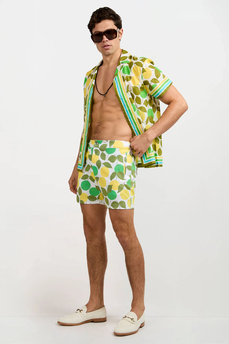 A man stands confidently against a white background wearing the Josue Short-Sleeve Shirt - Limone, matching leaf-print shorts, sunglasses, a necklace, and white loafers.