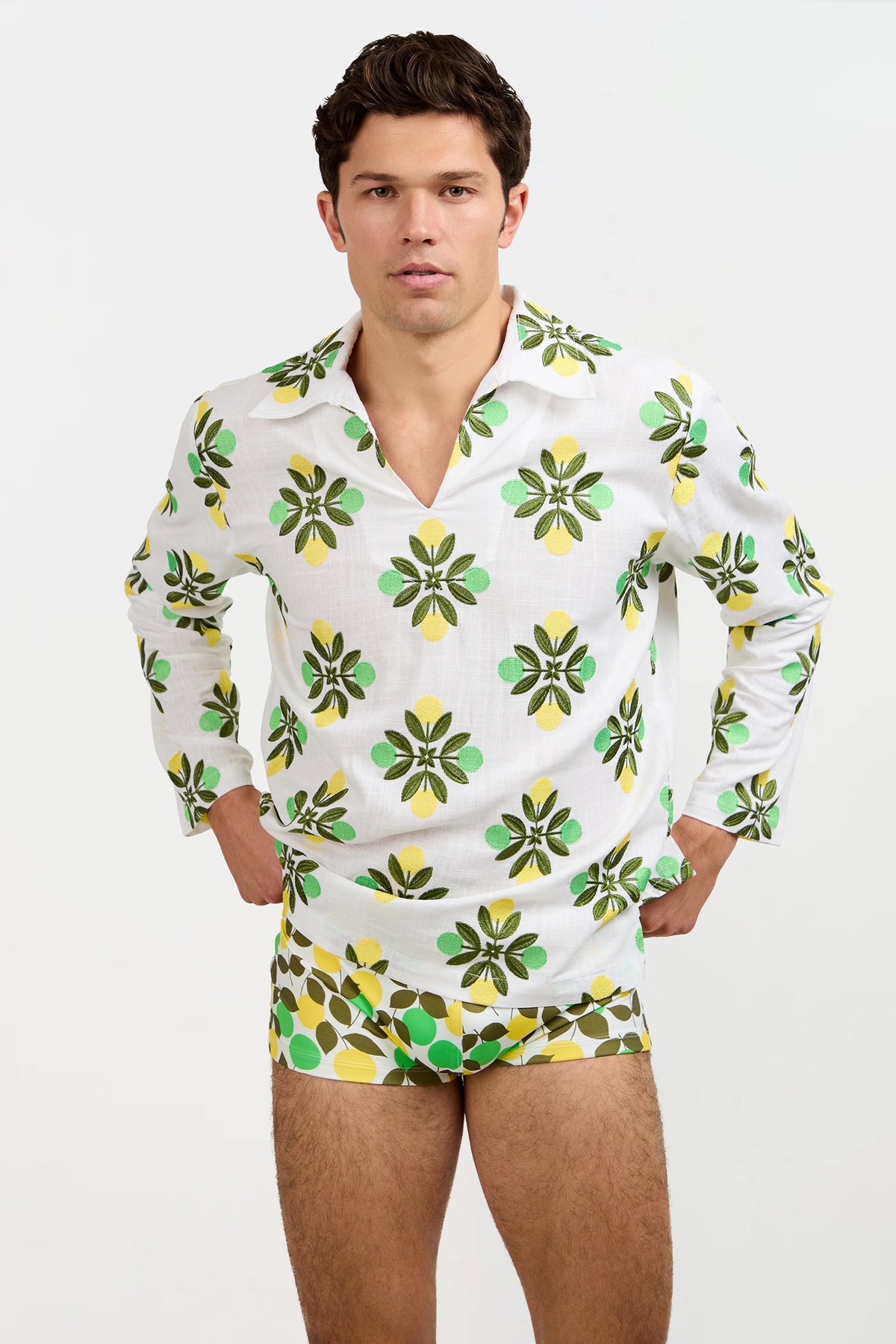 A man stands against a plain background, wearing the Holmes Pullover—a white long-sleeve shirt featuring a green and yellow floral pattern—with matching shorts. His hands rest on his hips as he looks directly at the camera.
