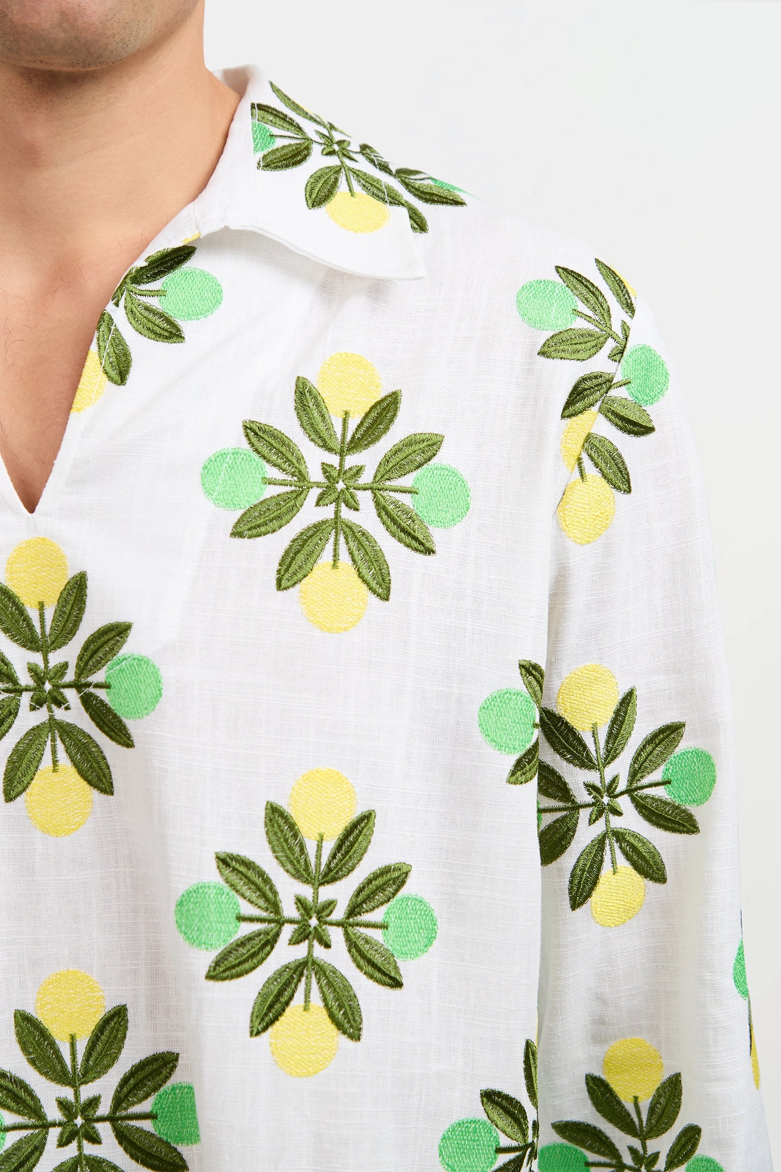 A close-up of the Holmes Pullover showcases its collared white fabric, adorned with embroidered green leaves and yellow-green circles, as seen on the upper torso and part of the wearer’s neck.