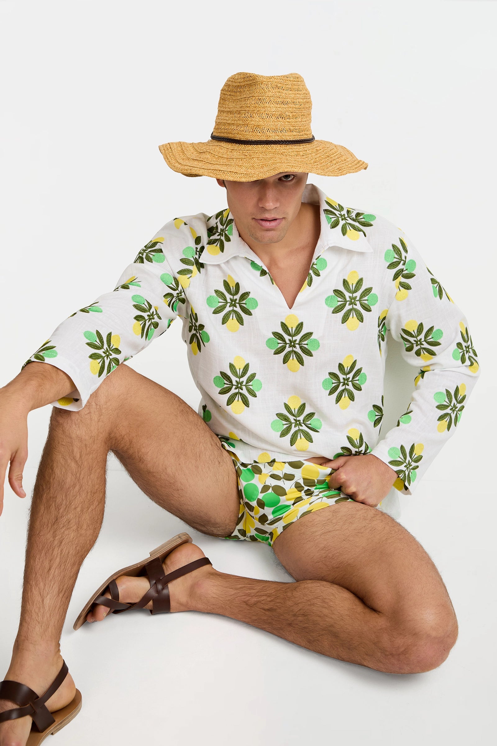 A person wearing the Holmes Pullover, patterned swim trunks, a straw sun hat, and brown sandals sits on the floor against a plain white background.