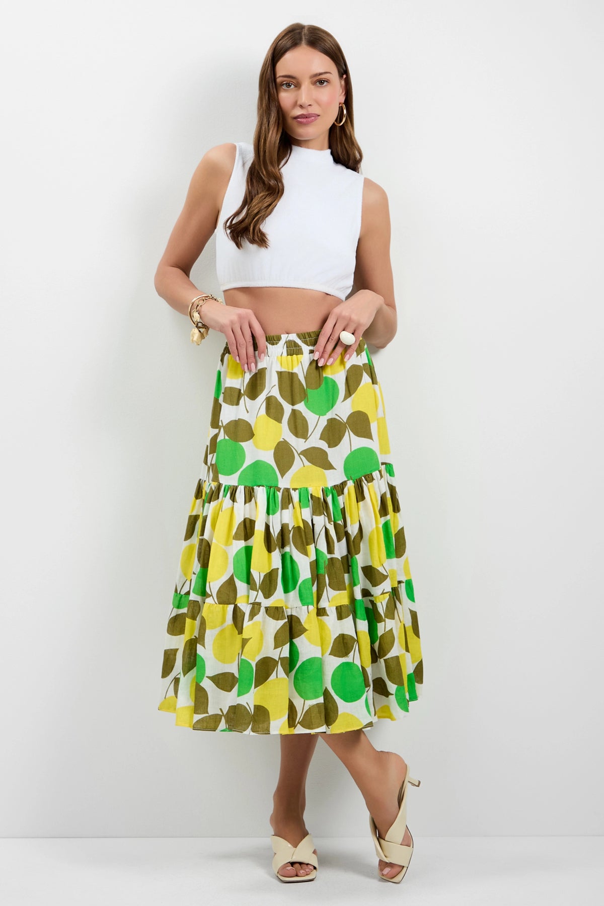 A woman stands against a white wall wearing a sleeveless white crop top, the Marsay Drop Waist Midi Skirt with a yellow, green, and brown circular pattern, beige heeled sandals, and gold bracelets.