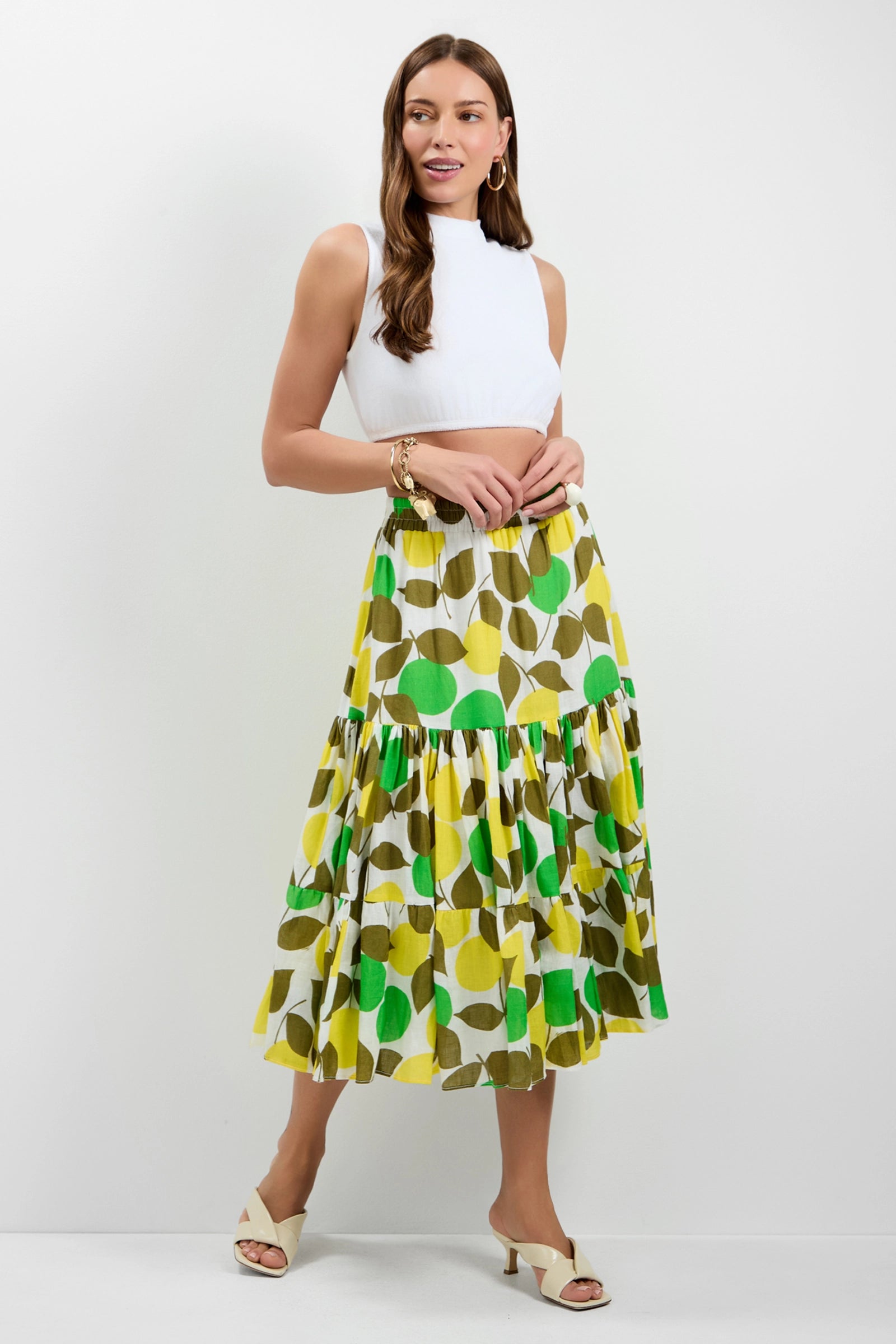 Woman in a sleeveless white crop top and the Marsay Drop Waist Midi Skirt, featuring a bold green, yellow, and brown circular print. She wears beige heeled sandals, her long brown hair down, smiling against a white background.