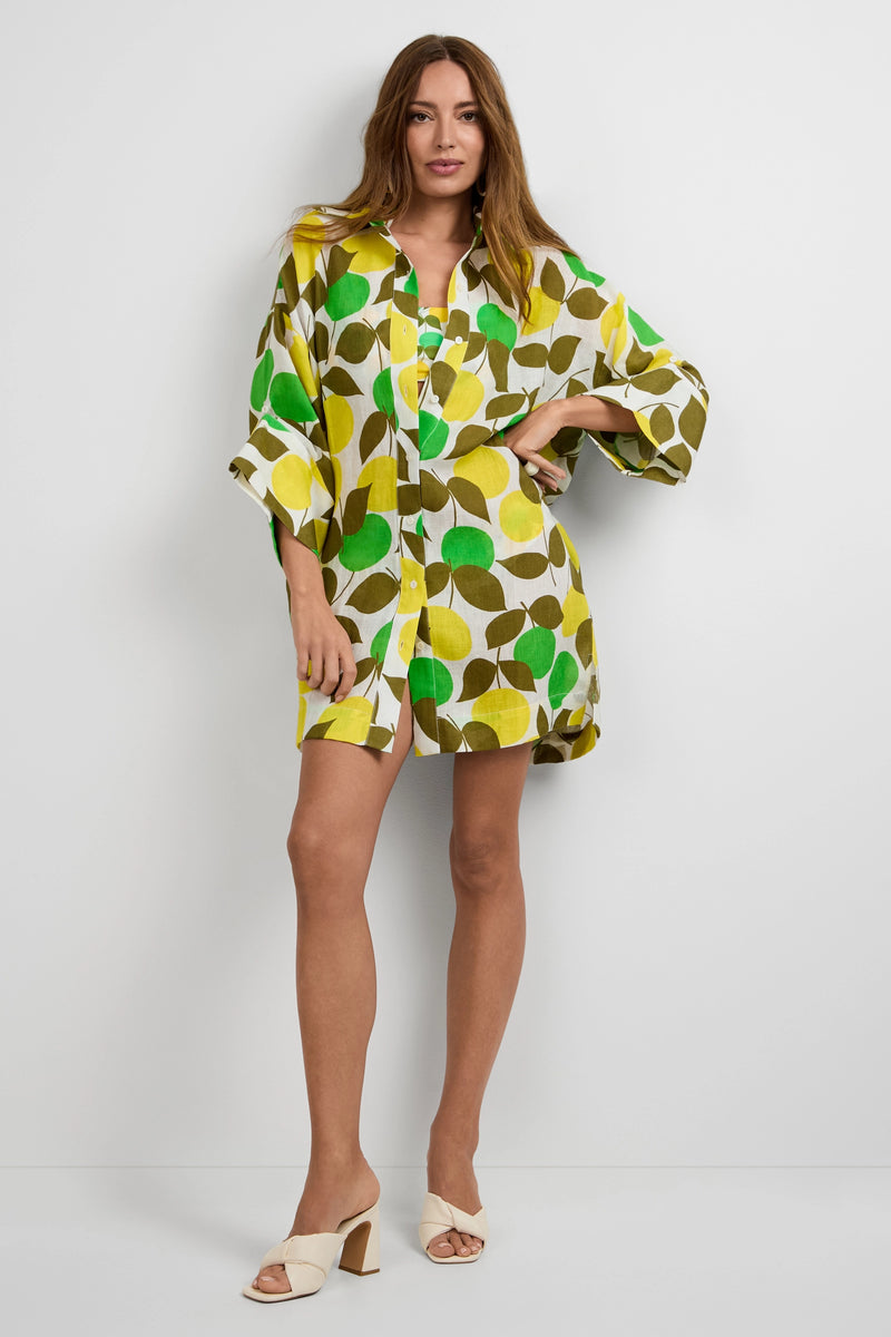 A woman with long brown hair stands against a white wall wearing the Opponent Shirt Dress featuring a bold green, yellow, and brown leaf print, paired with cream heeled sandals.