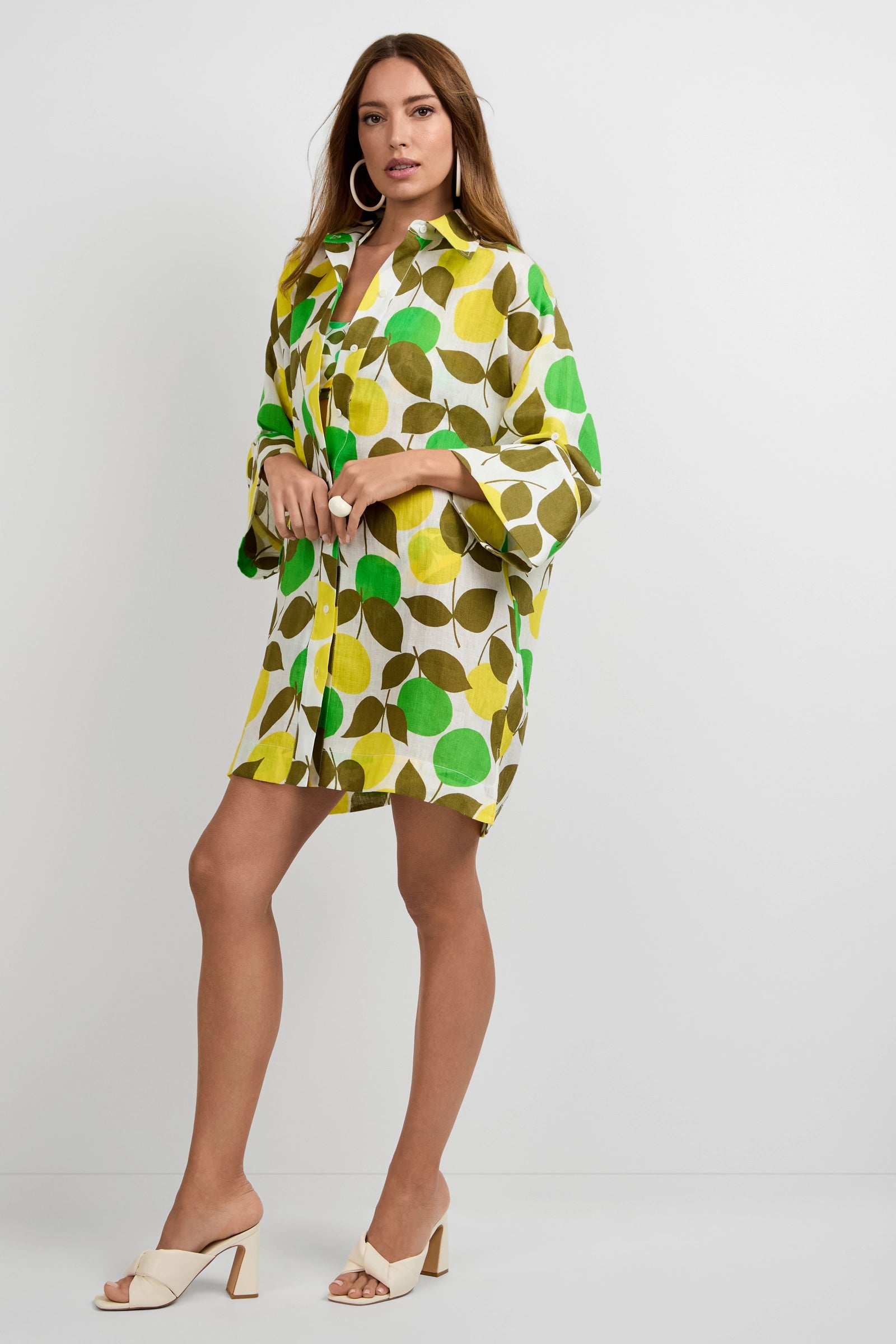 A woman wearing the Opponent Shirt Dress with a green, yellow, and brown leaf pattern stands against a plain background. She has long hair, large hoop earrings, and white heeled sandals.