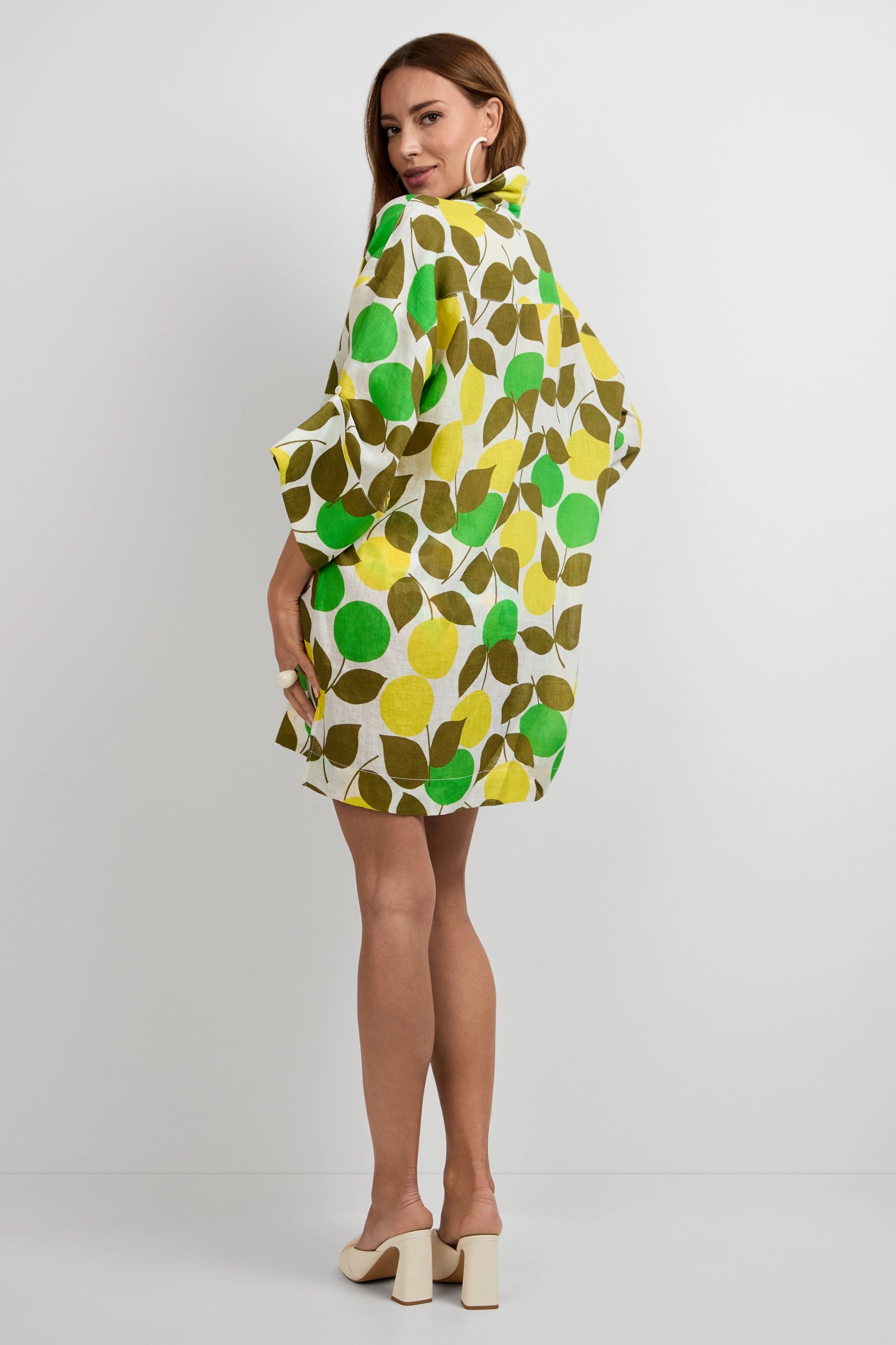 A woman with long hair, facing away, wears the Opponent Shirt Dress—a loose, knee-length piece with green, yellow, and brown leaf patterns—paired with cream heeled sandals against a plain white background.