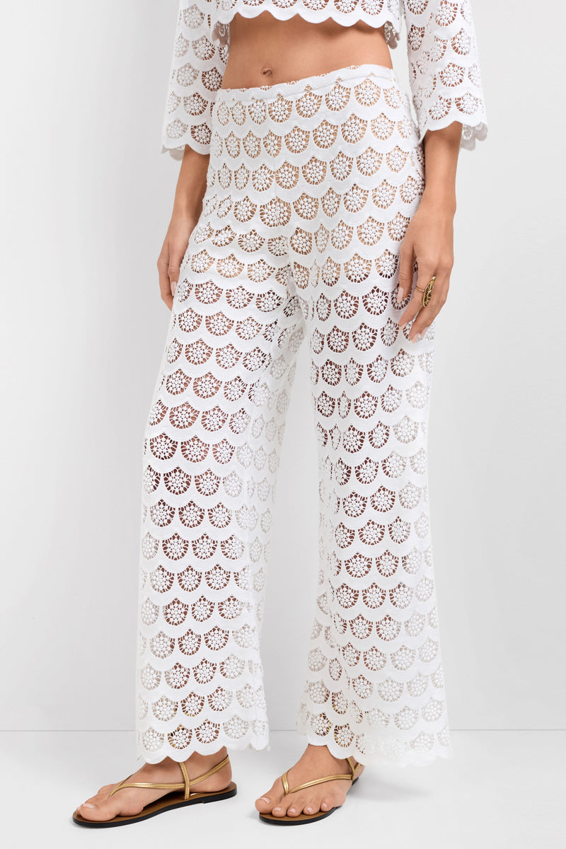 A person in semi-sheer Dzo Dzo Crochet Lace Pants with a scalloped pattern, paired with a matching cropped top and gold sandals. The wide-leg, high-waisted pants and top both reveal skin beneath.