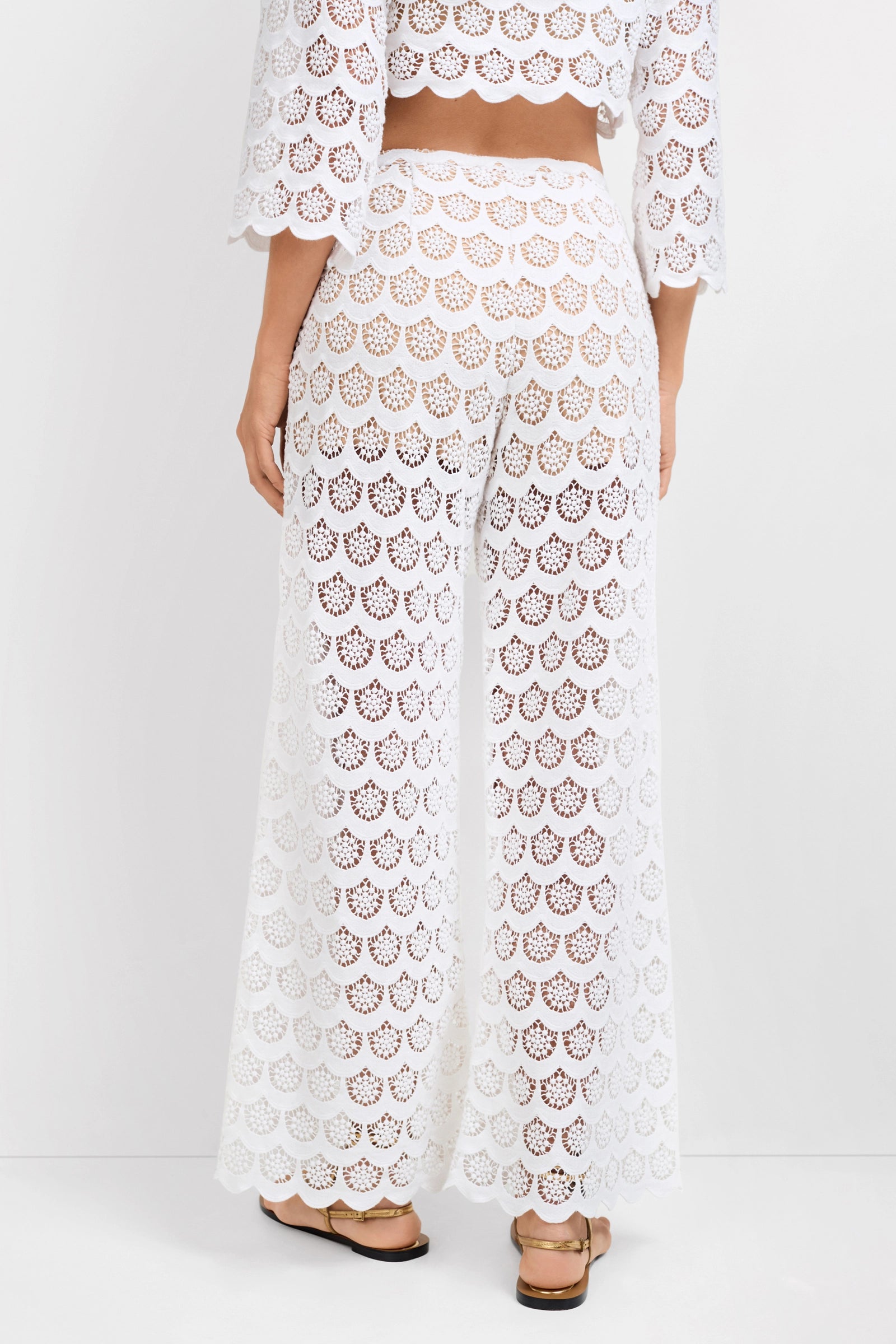 A person stands facing away, wearing the semi-sheer Dzo Dzo Crochet Lace Pants with a matching scalloped lace top and sandals. The background is plain white.
