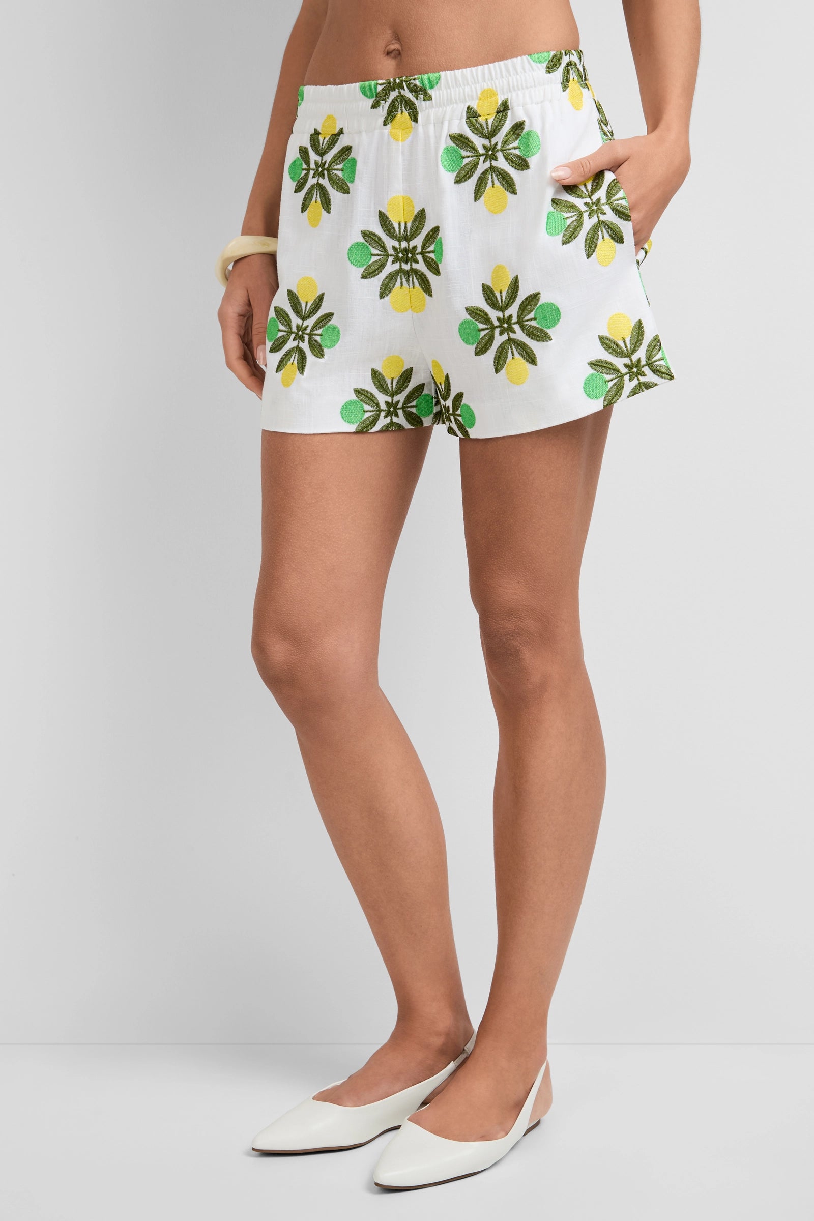 A person wearing Azule Shorts with green and yellow floral prints stands against a plain background, hands in pockets. The look is styled with a white bracelet and white pointed-toe flats.