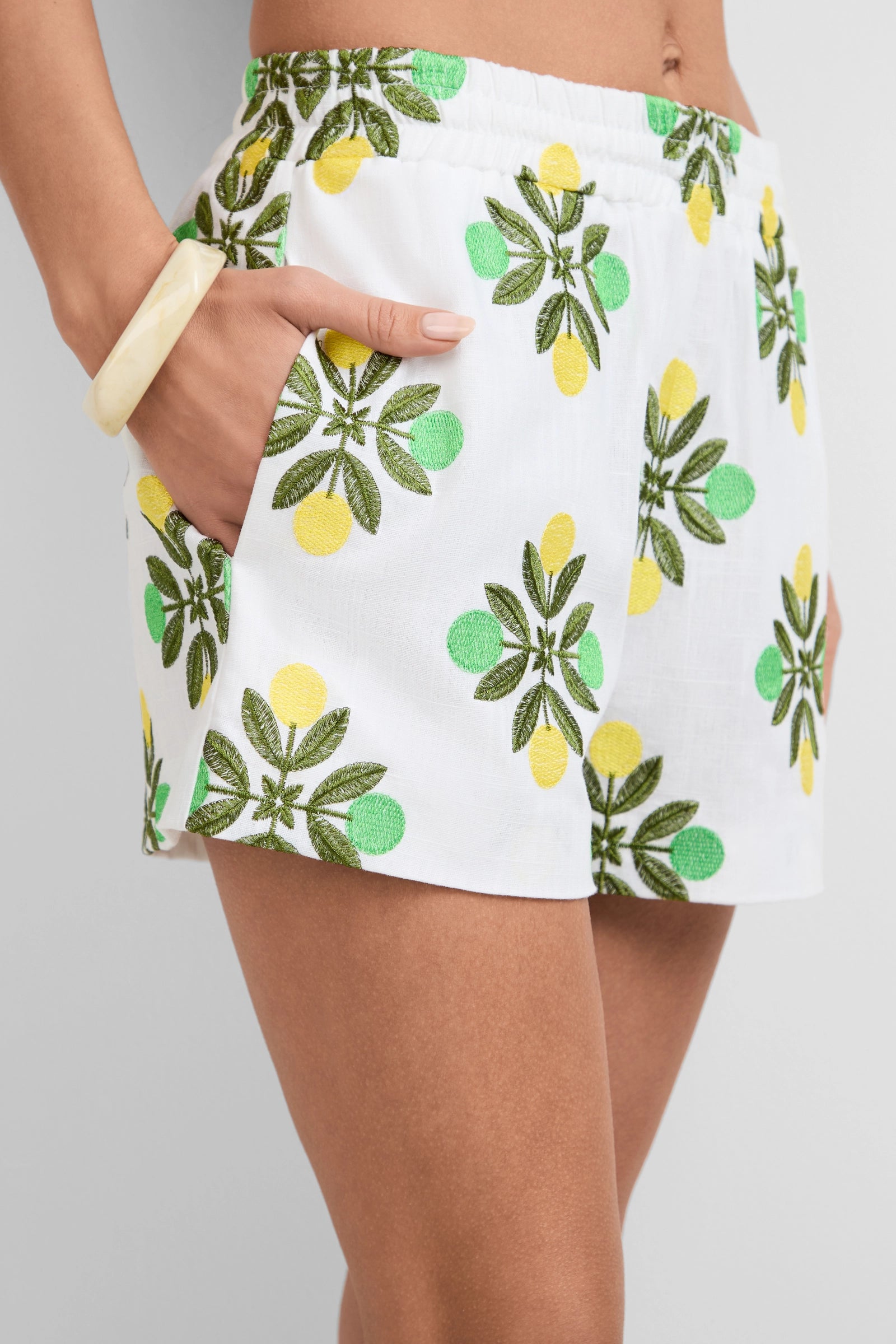 Someone wearing the Azule Shorts, which feature white fabric with green and yellow lemon leaf patterns, stands with one hand in their pocket and a chunky light-colored bracelet on their wrist.