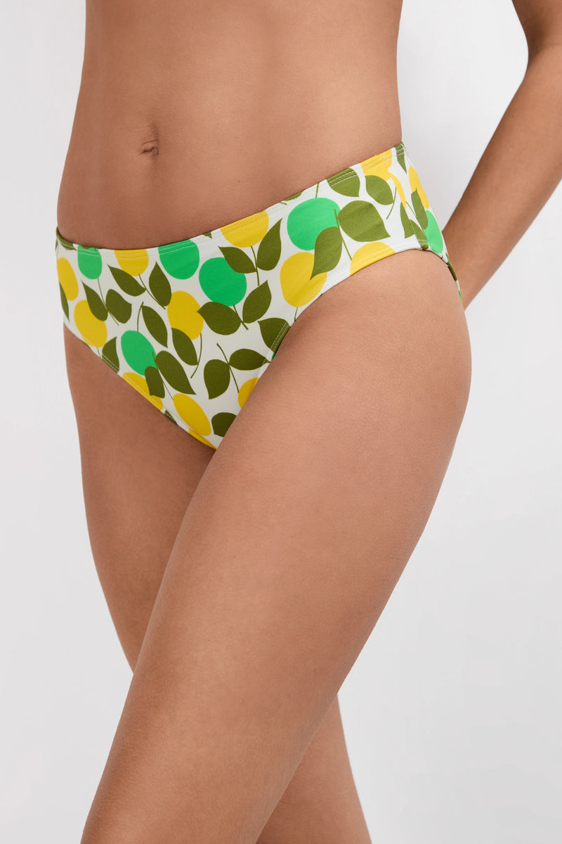 A person wears the Arissa Hipster Swim Bottom - Limone, featuring a green, yellow, and olive leaf pattern on white; only their torso and upper thighs are visible against a plain background.