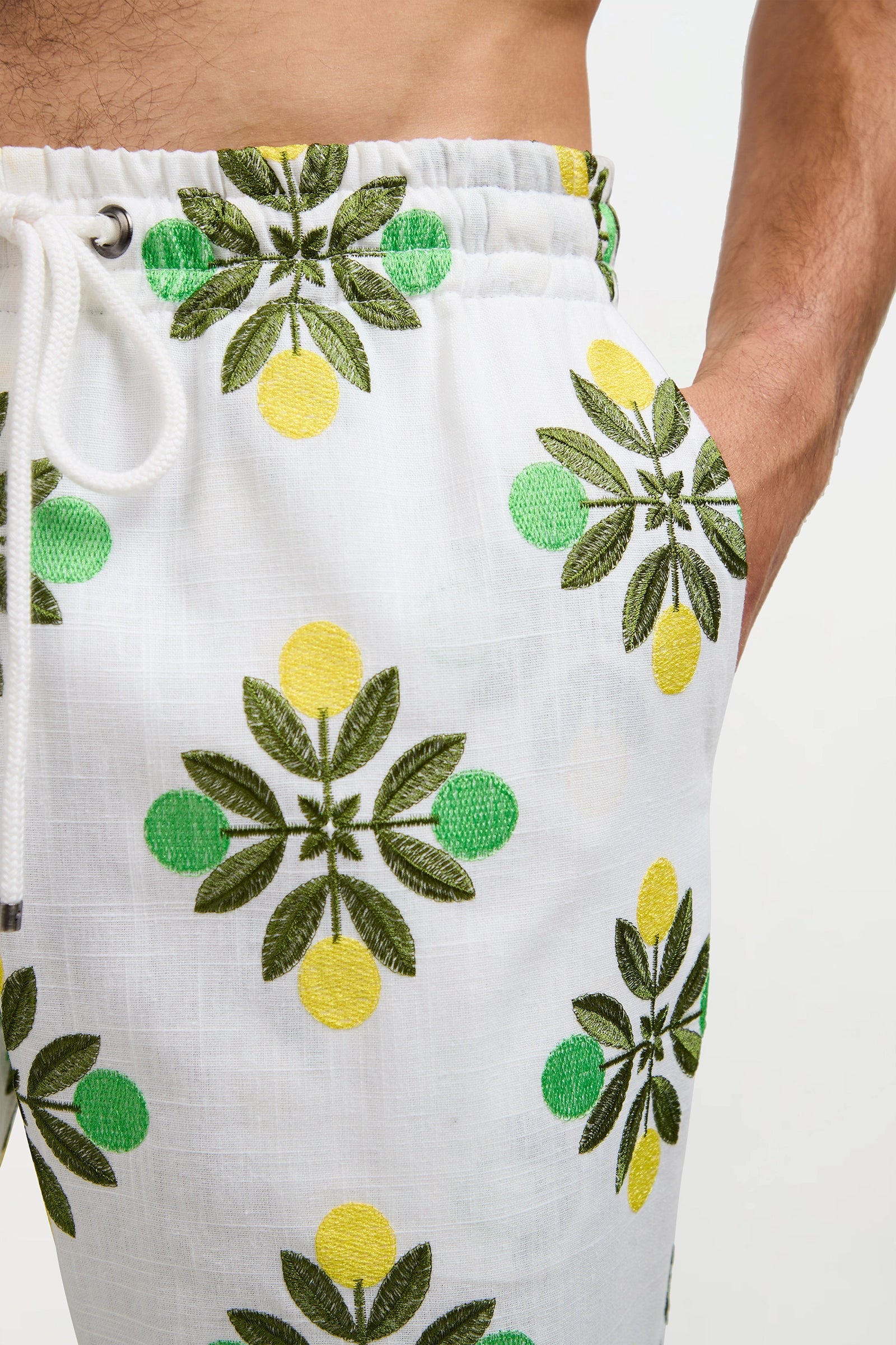 A close-up of a person wearing the Felipe Drawstring Pant, featuring white fabric with a green leaf and yellow lemon pattern, an elastic waistband with drawstring, and a hand in the pocket.