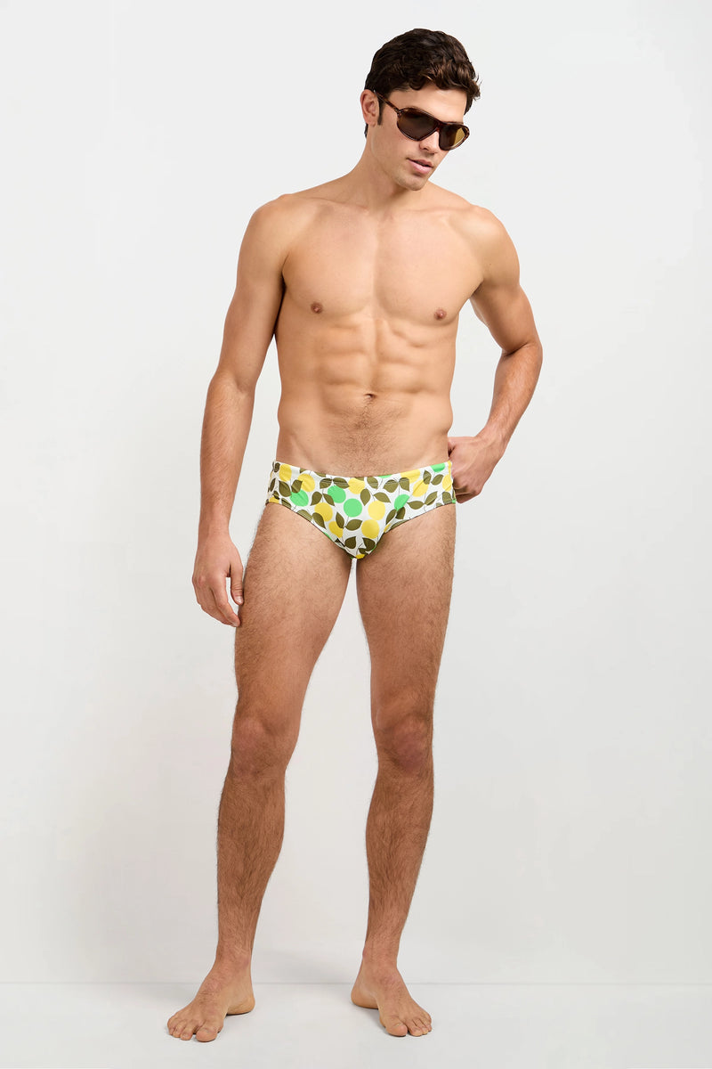 A man models the Bondi Swim Brief - Limone and sunglasses, standing barefoot against a plain white background and looking downward with one arm by his side.