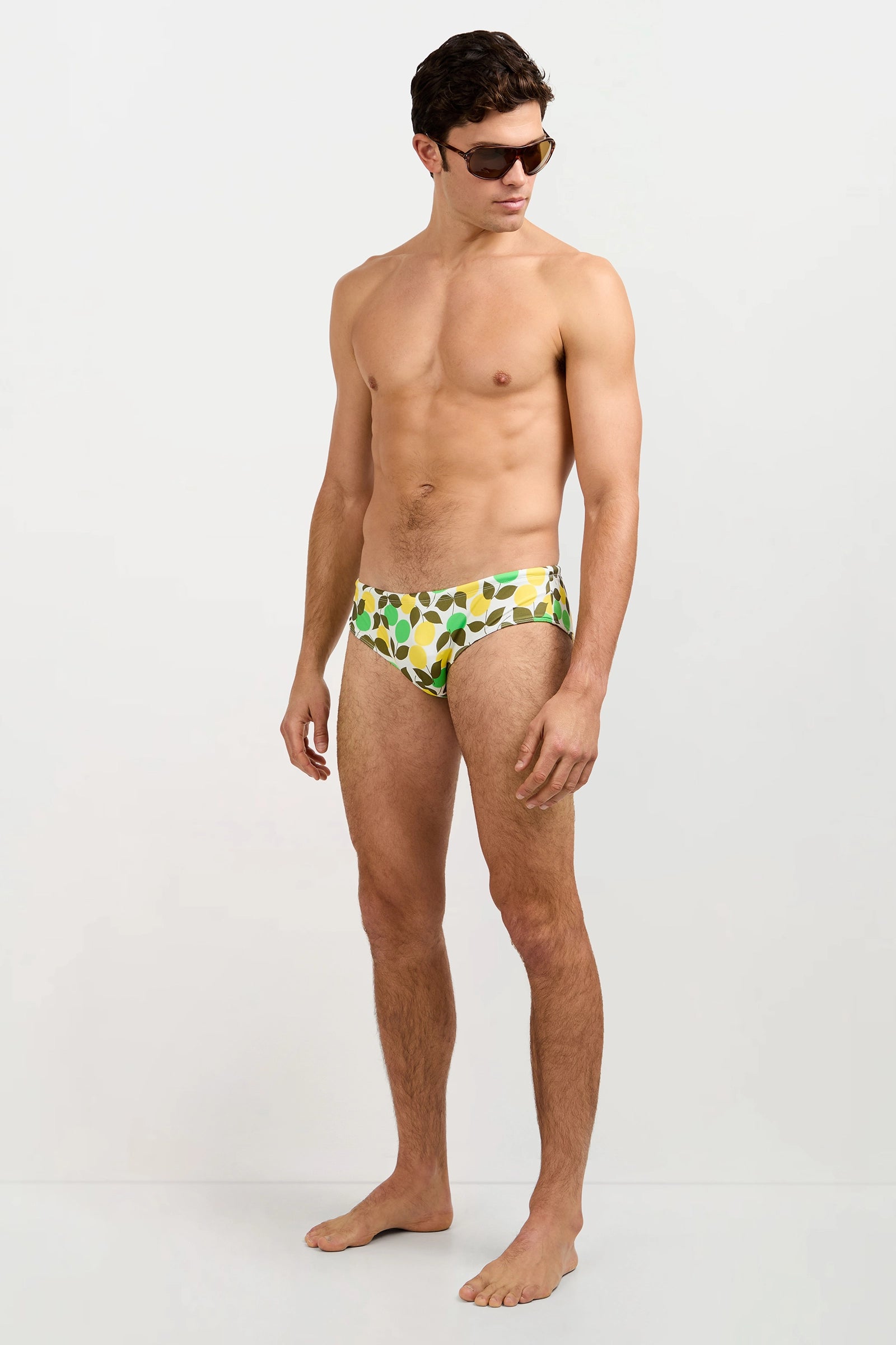 A man models the Bondi Swim Brief - Limone in a yellow-green pattern, barefoot and wearing sunglasses, standing against a plain white background and looking down to his left.