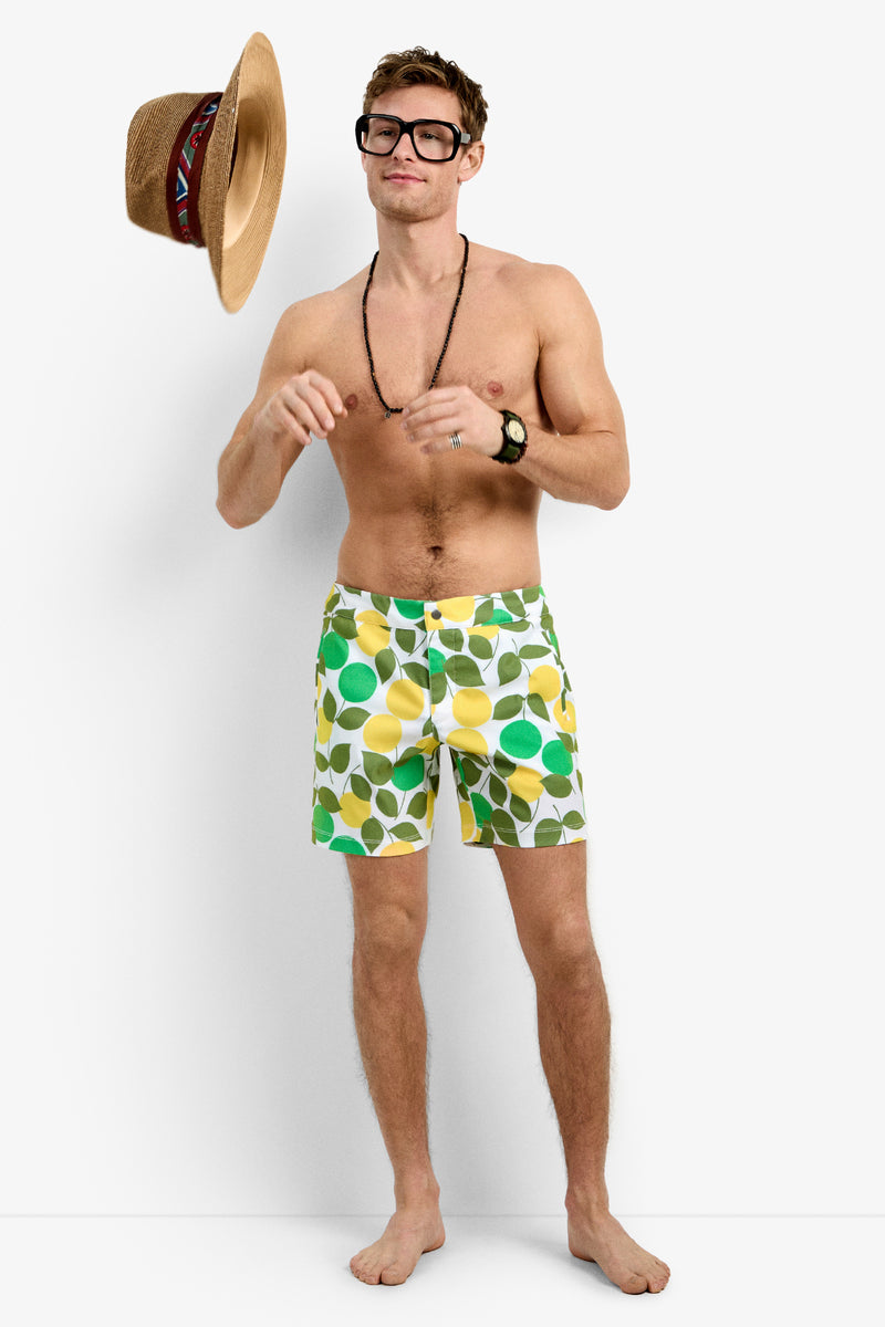 A smiling, shirtless man in glasses, a necklace, a wristwatch, and Honolulu Board Short tosses a straw hat into the air against a plain white background.