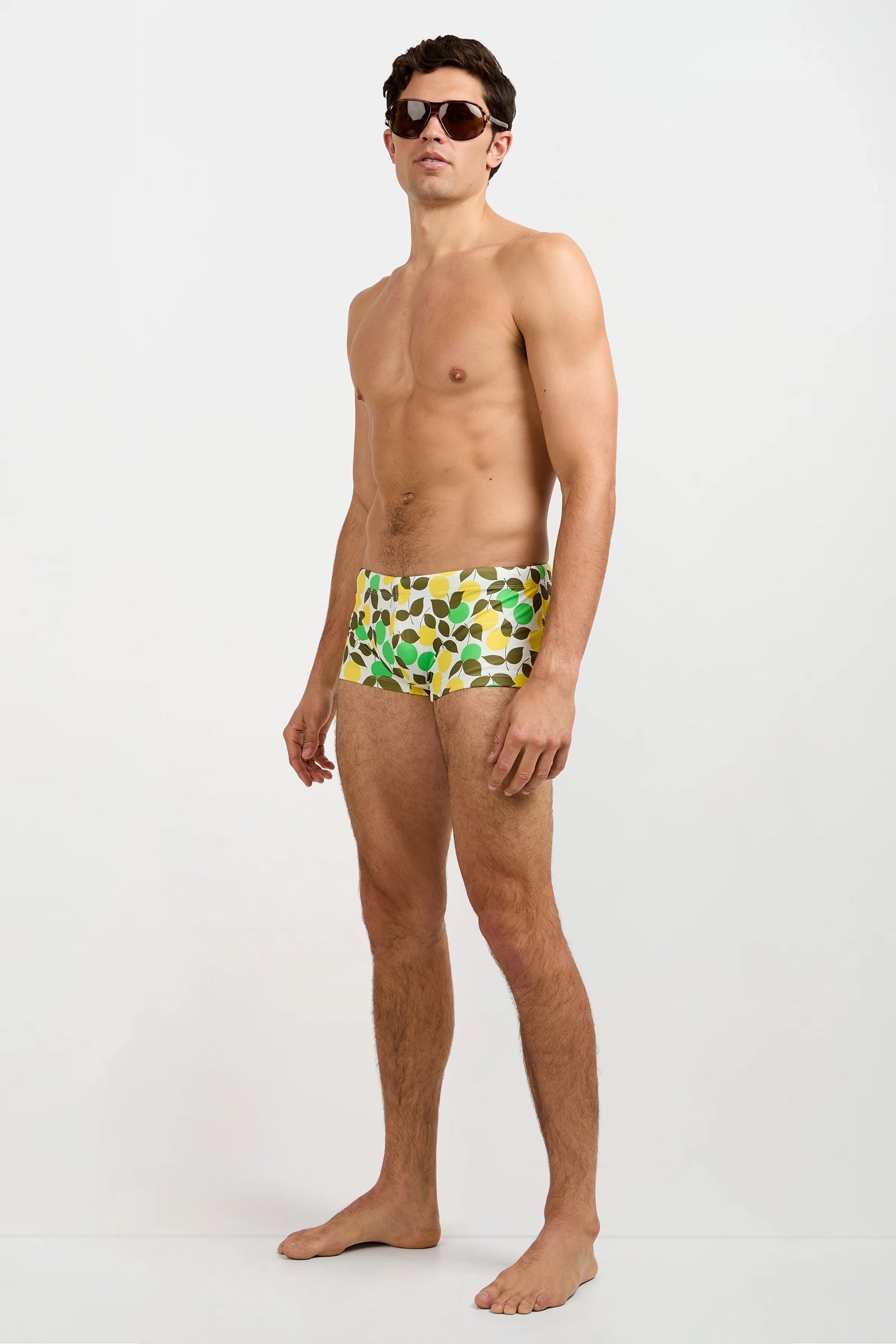 A man wearing dark sunglasses and the Sorrento Swim Trunk - Limone stands barefoot against a plain white background.